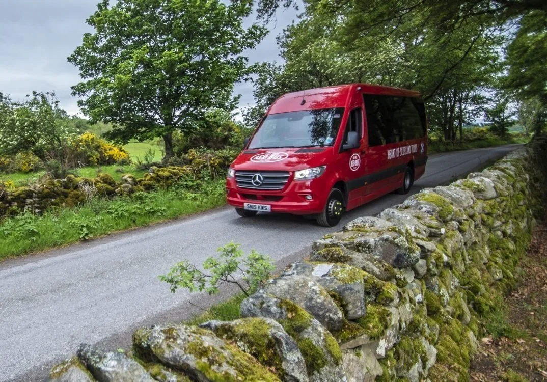 Red Mercedes-Benz minibus with Coca-Cola logo and promotional writing on a rural road next to a moss-covered stone wall, surrounded by green trees and shrubs.