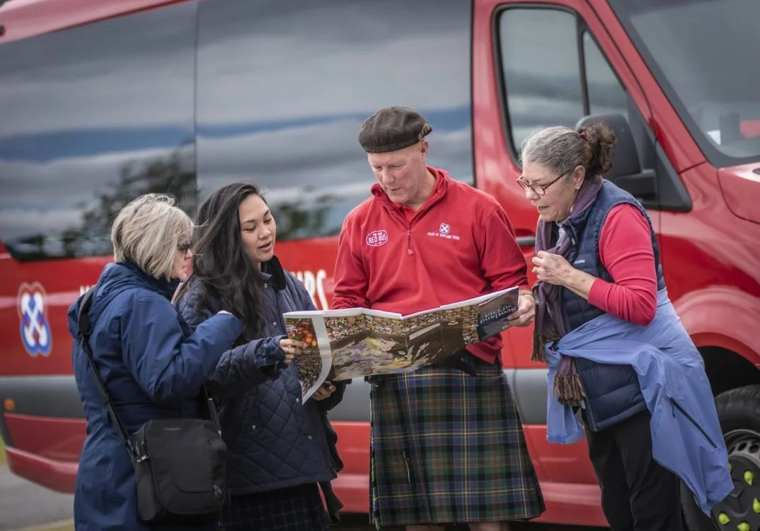 Four people, two women, a man, and a girl, are gathered outdoors in front of a red tour bus, looking at a map.