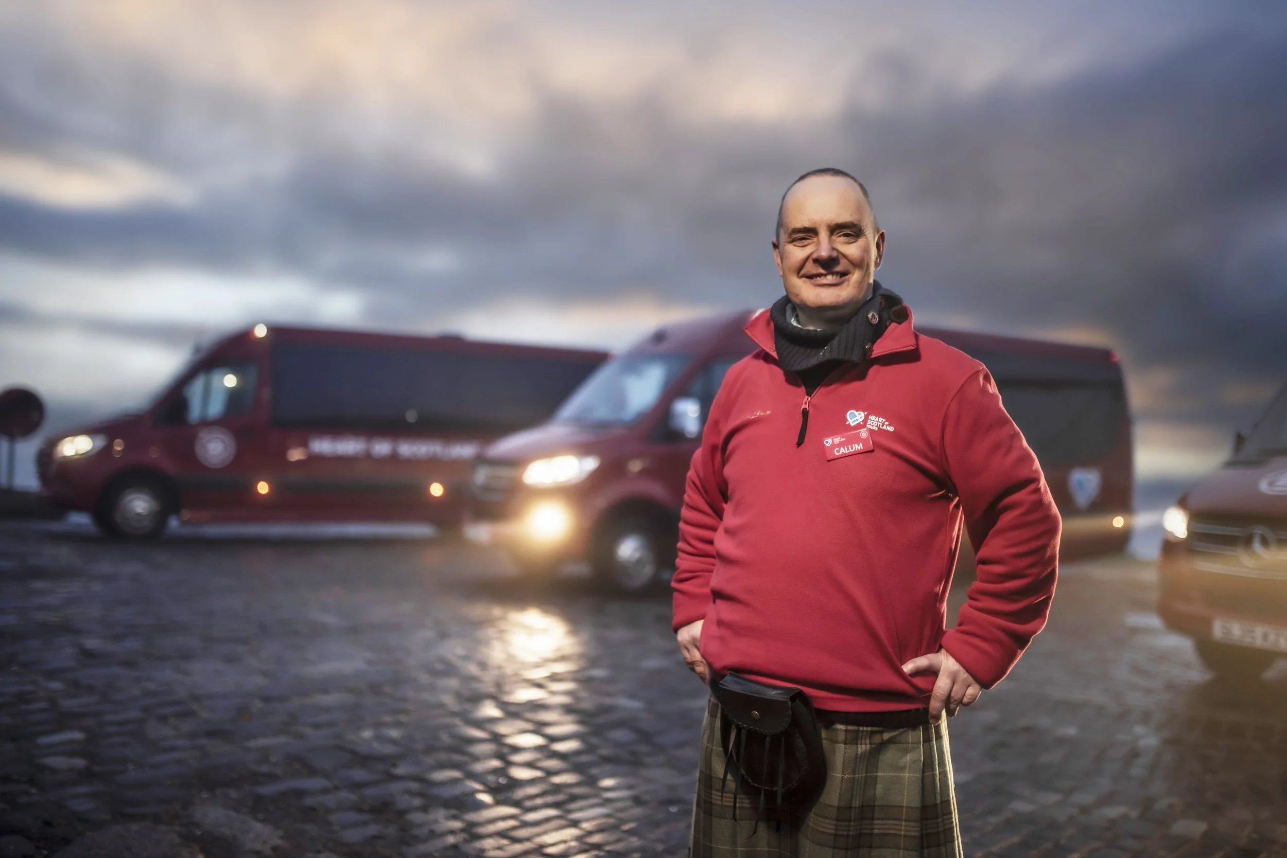 A smiling man wearing a red jacket and plaid pants standing outdoors on a rainy day with dark clouds in the sky and red vans in the background.