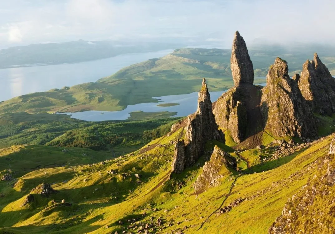 Tall, jagged rock formations on a lush, green hillside overlooking lakes and distant mountains under a partly cloudy sky.
