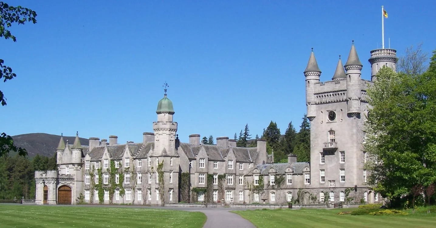 A large, gray castle with multiple turrets and towers, surrounded by a well-maintained lawn and trees, under a clear blue sky.