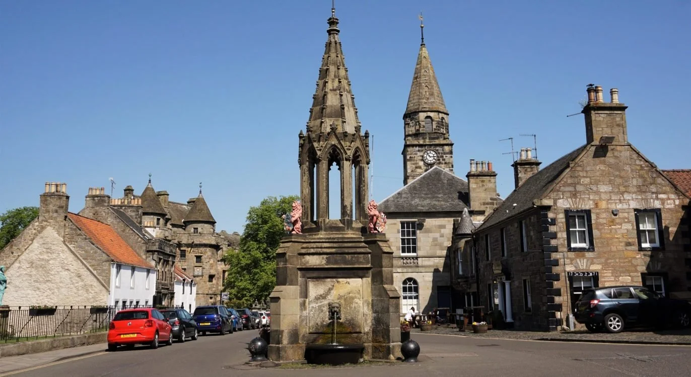 A street scene in a European town with historic stone buildings, a decorative stone fountain with pink lion sculptures, and a church with a tall spire and clock tower under a clear blue sky.