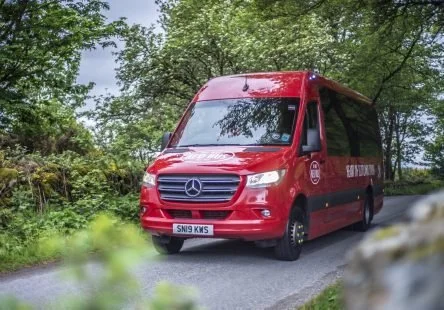 Red Mercedes-Benz van with district logo driving on a rural road surrounded by trees.