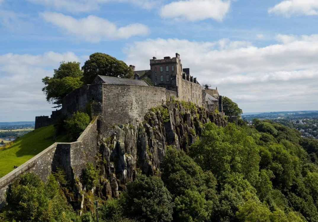 A historic castle on a hilltop overlooking a lush green landscape and distant town under a partly cloudy sky.