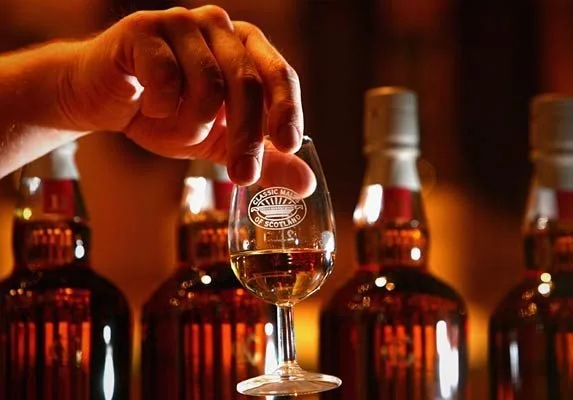A hand holding a small glass of scotch, with bottles of whiskey in the background.