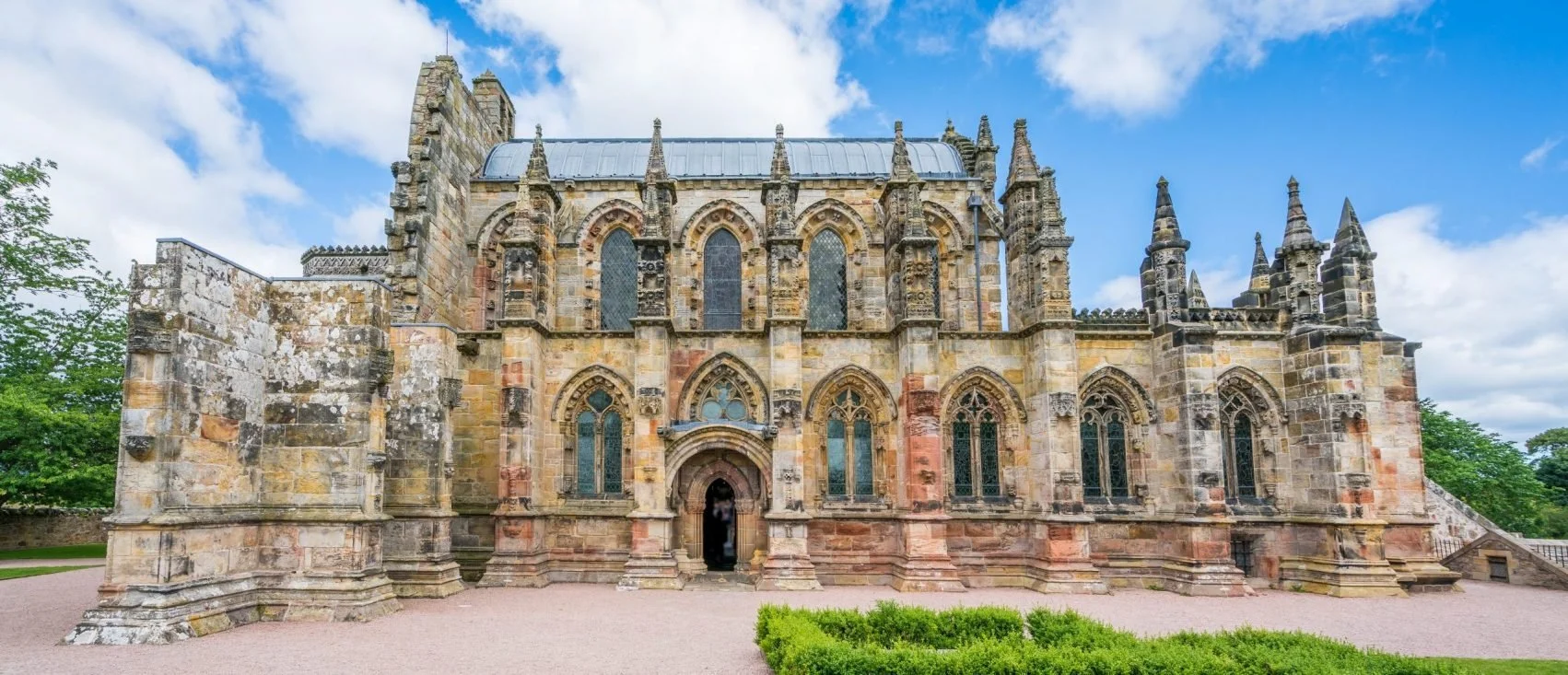 Photo of a historic Gothic-style stone church with pointed arches and tall, narrow stained glass windows, surrounded by a gravel courtyard and some green trees in the background.