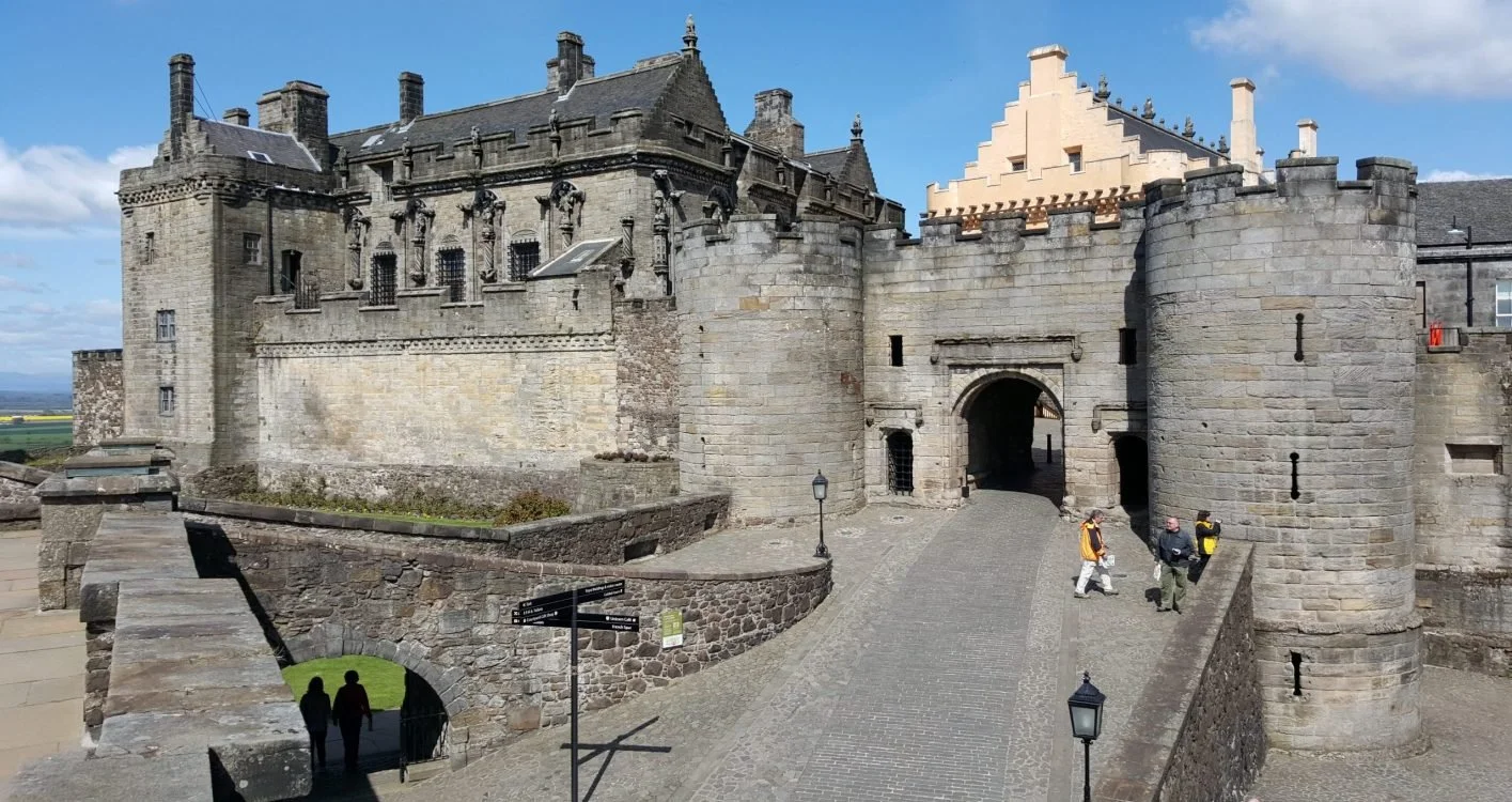 View of Stirling castle with battlements and towers, showing an arched entryway, cobblestone pathway, and a few tourists walking near the entrance, under a partly cloudy sky.