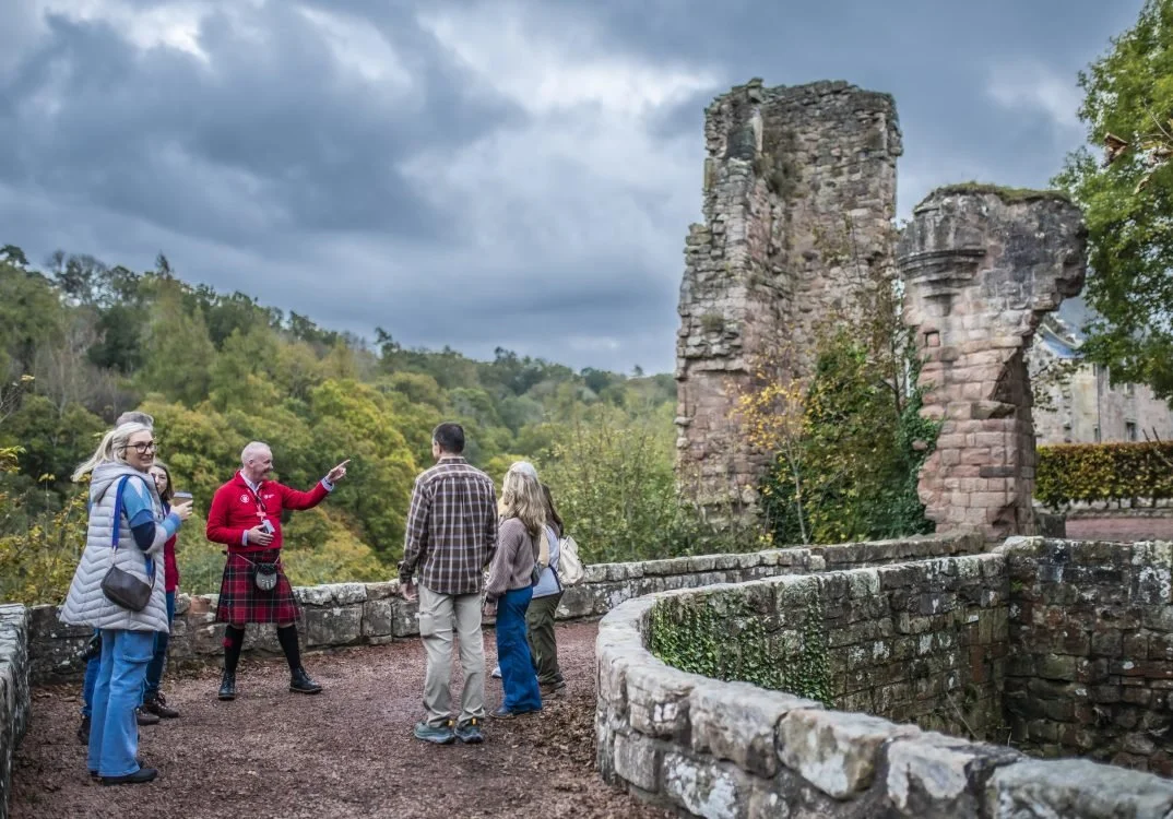 Tour guide in traditional Scottish attire speaking to a group of tourists at a historic site with stone ruins and a cloudy sky.