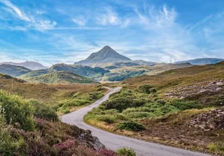 A dirt road winding through a hilly landscape with green and purple vegetation, leading toward a mountain in the background under a blue sky with wispy clouds.