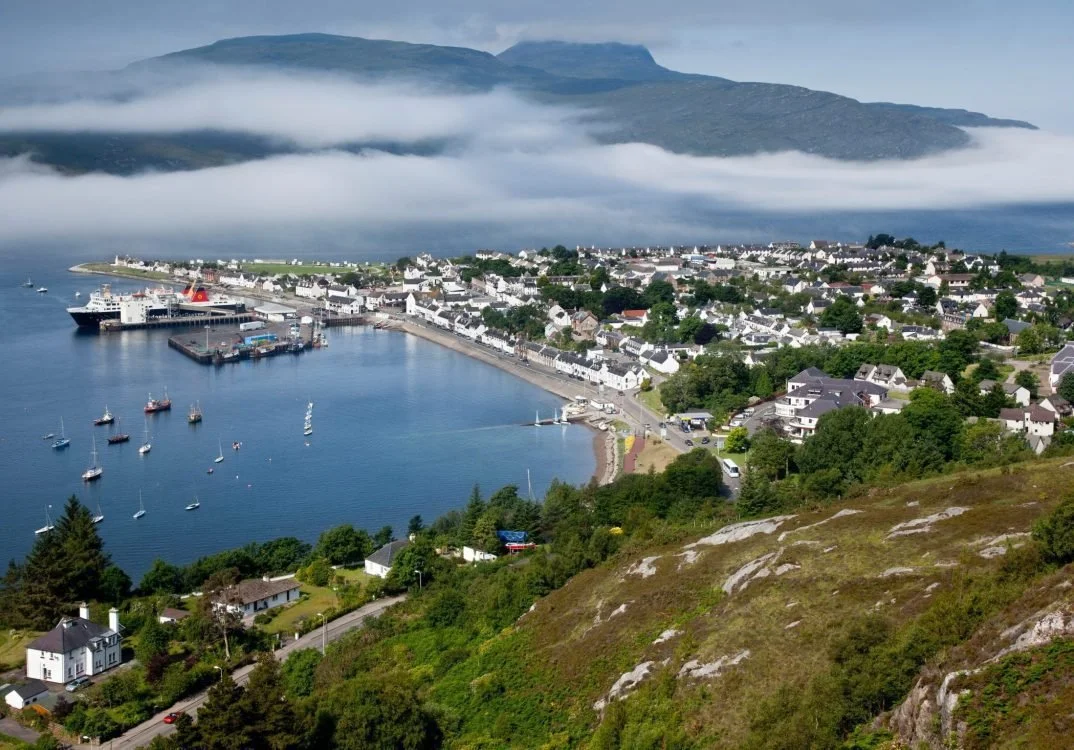 A coastal town with white buildings, a harbor with boats, surrounded by lush green hills, and mountains in the background partially covered by clouds.