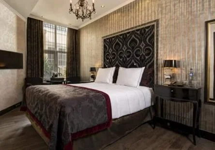 A well-decorated hotel room with a large bed, patterned headboard, bedside tables with lamps, and windows with curtains.