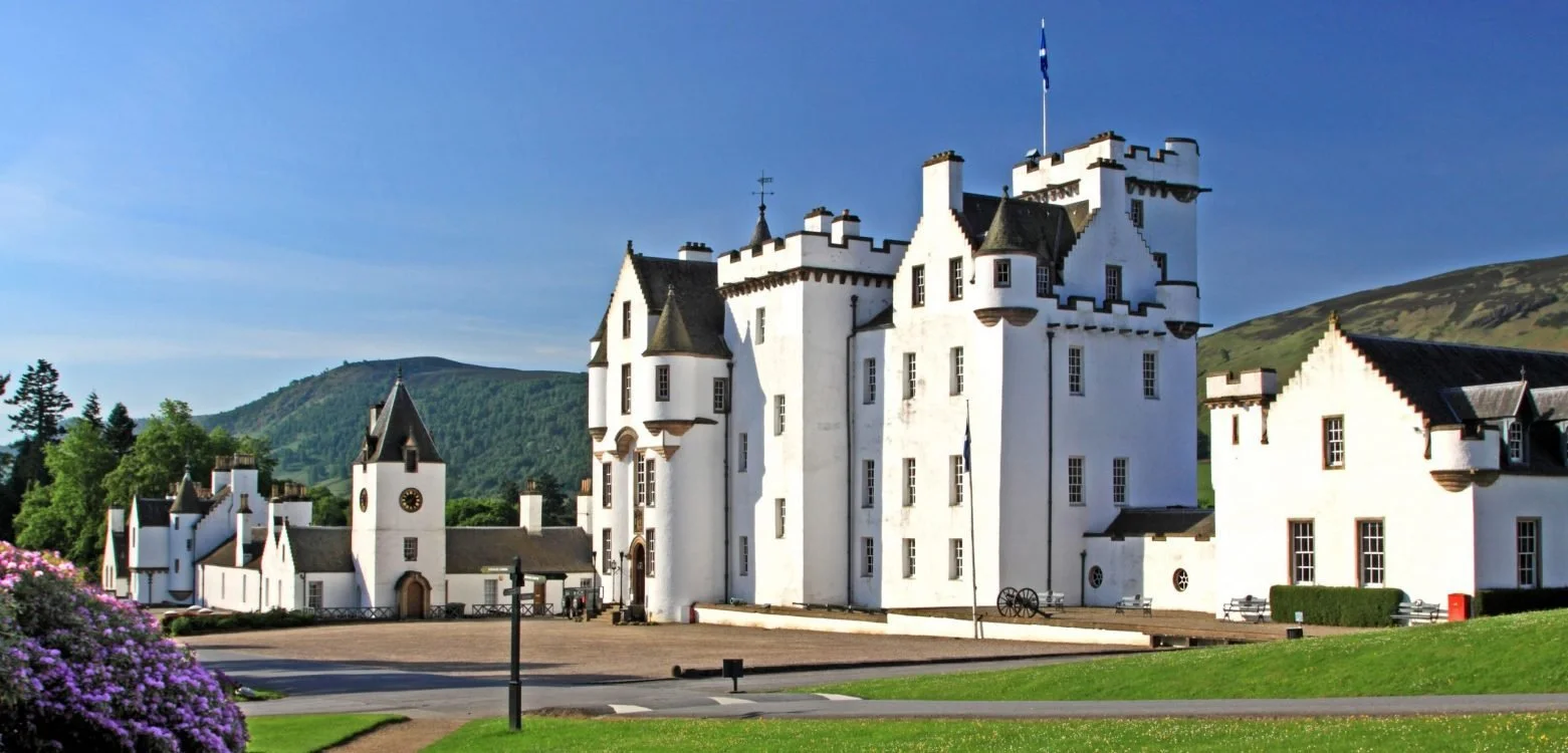 A large white castle with black rooftops and turrets, surrounded by greenery and mountains in the background, under a clear blue sky.