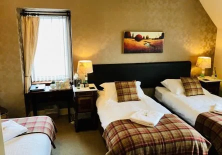 A hotel bedroom with two double beds, a window with curtains, and two bedside lamps. The beds have plaid blankets and pillows.