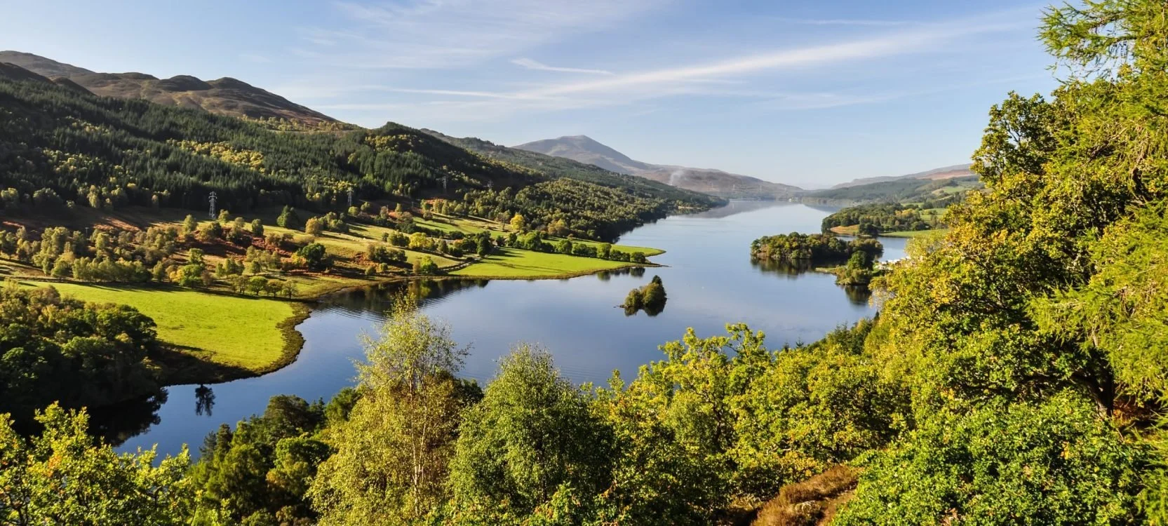 A scenic view of a river flowing through green hills and trees with mountains in the background under a clear blue sky.