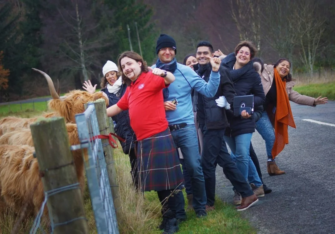 Group of six diverse people smiling and posing along a rural road with Highland cows, surrounded by trees and grassy fields.