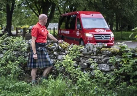 Man in a red shirt and kilt leaning on a stone wall in a park-like setting with trees and a red van parked behind him.