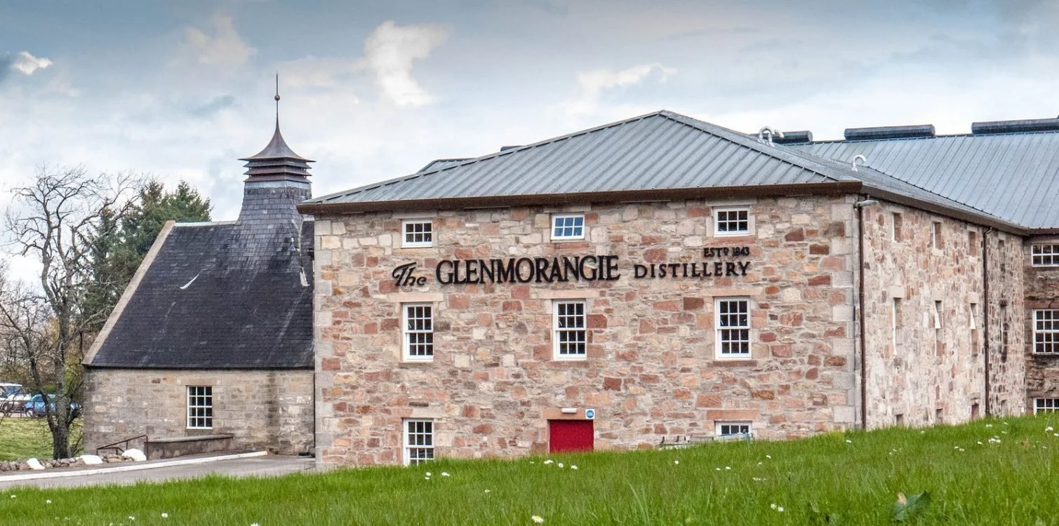 A stone building with a sign that reads "The Glenmorangie Distillery" and has small white-framed windows, set against a backdrop of cloudy sky and some trees, with a grassy foreground.