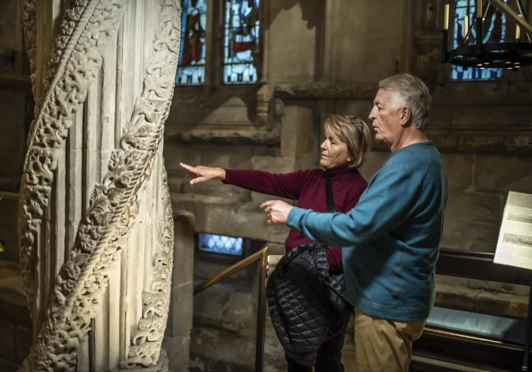 Two elderly people, a man and a woman, observing and discussing an intricately carved stone pillar inside a historic building, with stained glass windows in the background.