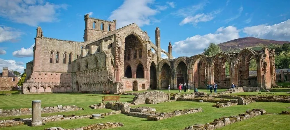 Ruins of an ancient stone church or abbey with arched windows and a grassy foreground under a partly cloudy sky.