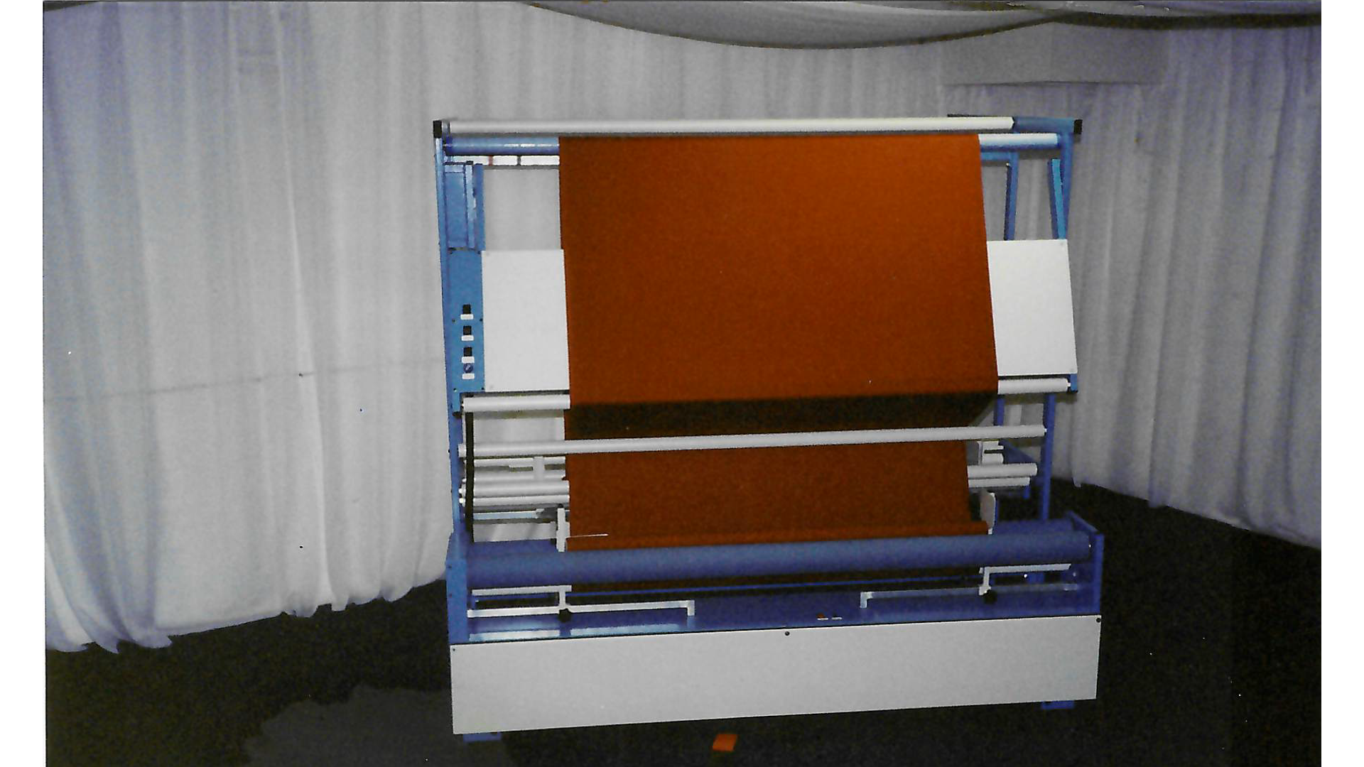 A rolling machine with a blue metal frame, white rollers, and orange fabric or material, set against white curtains.