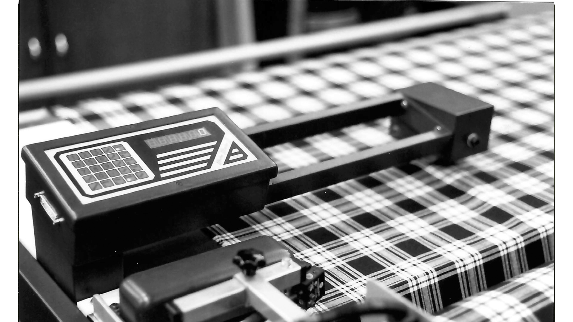 Electronic device with a small screen and keypad placed on a checkered tablecloth, with metallic machinery in the background.