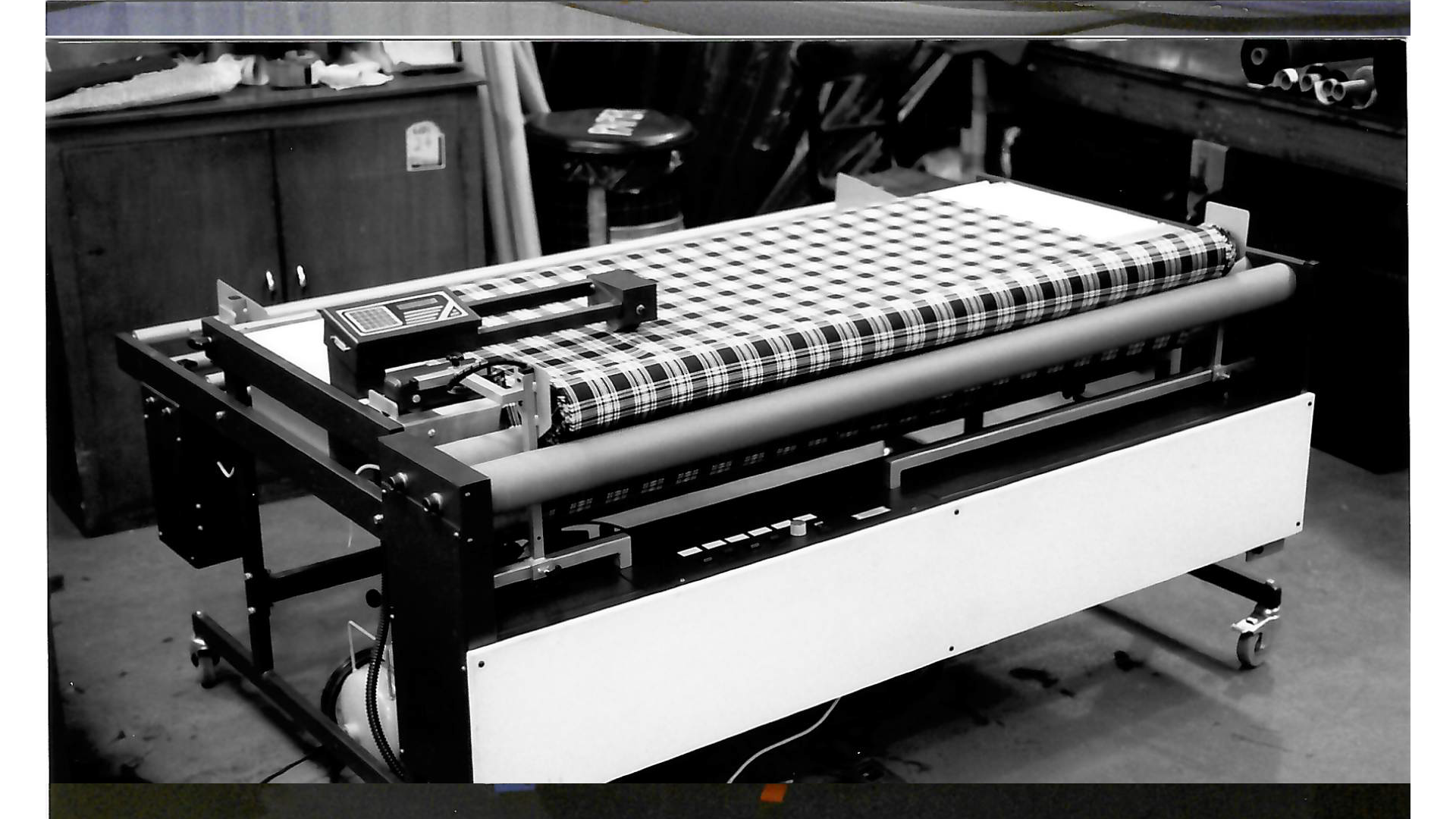 A cutting machine with a plaid fabric on a worktable in a workshop or factory setting.