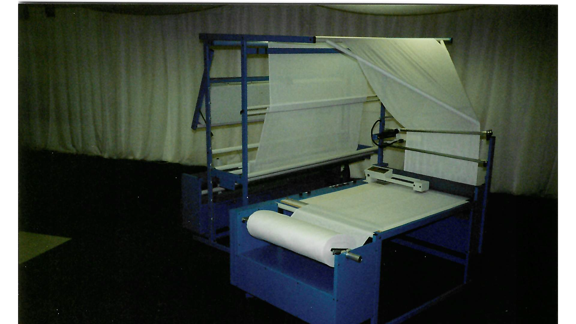 A large blue and white fabric cutting machine with a rolling fabric roll, set up indoors against a plain curtain background.