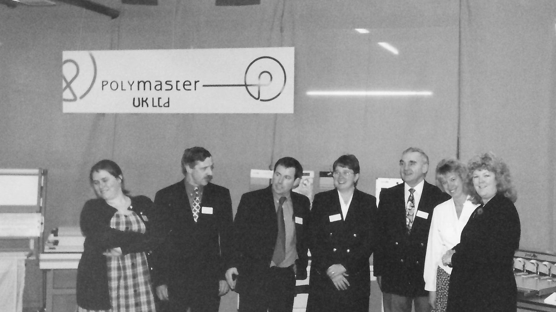 Black and white photo of six people in business attire standing in line, smiling, with a sign overhead that says 'Polymaster UK Ltd.' and a diagram of a circular object above the text.