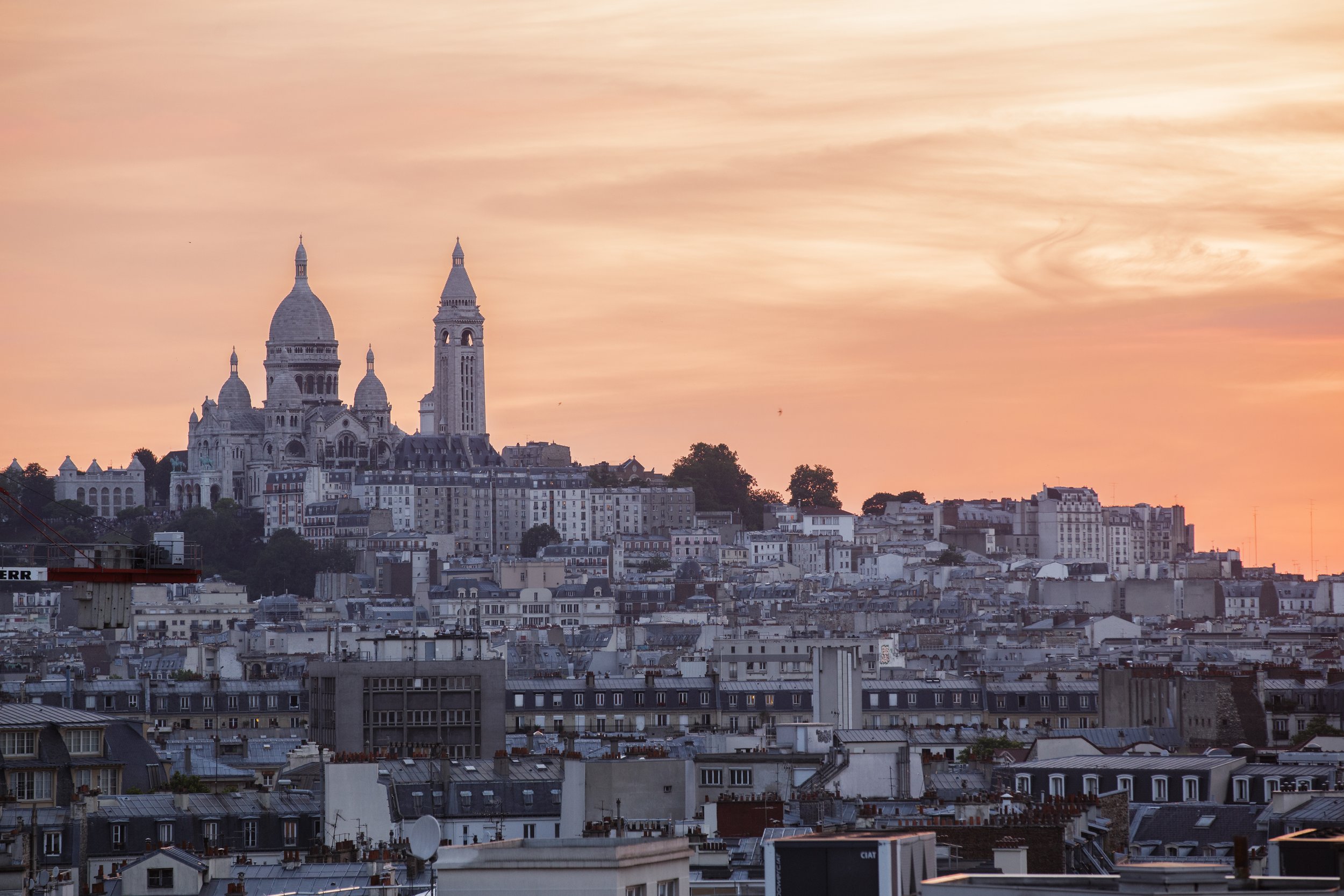 A panoramic view of Paris at sunset, featuring the Sacré-Cœur Basilica with its distinctive domes on the hilltop and the surrounding cityscape with various buildings and rooftops.