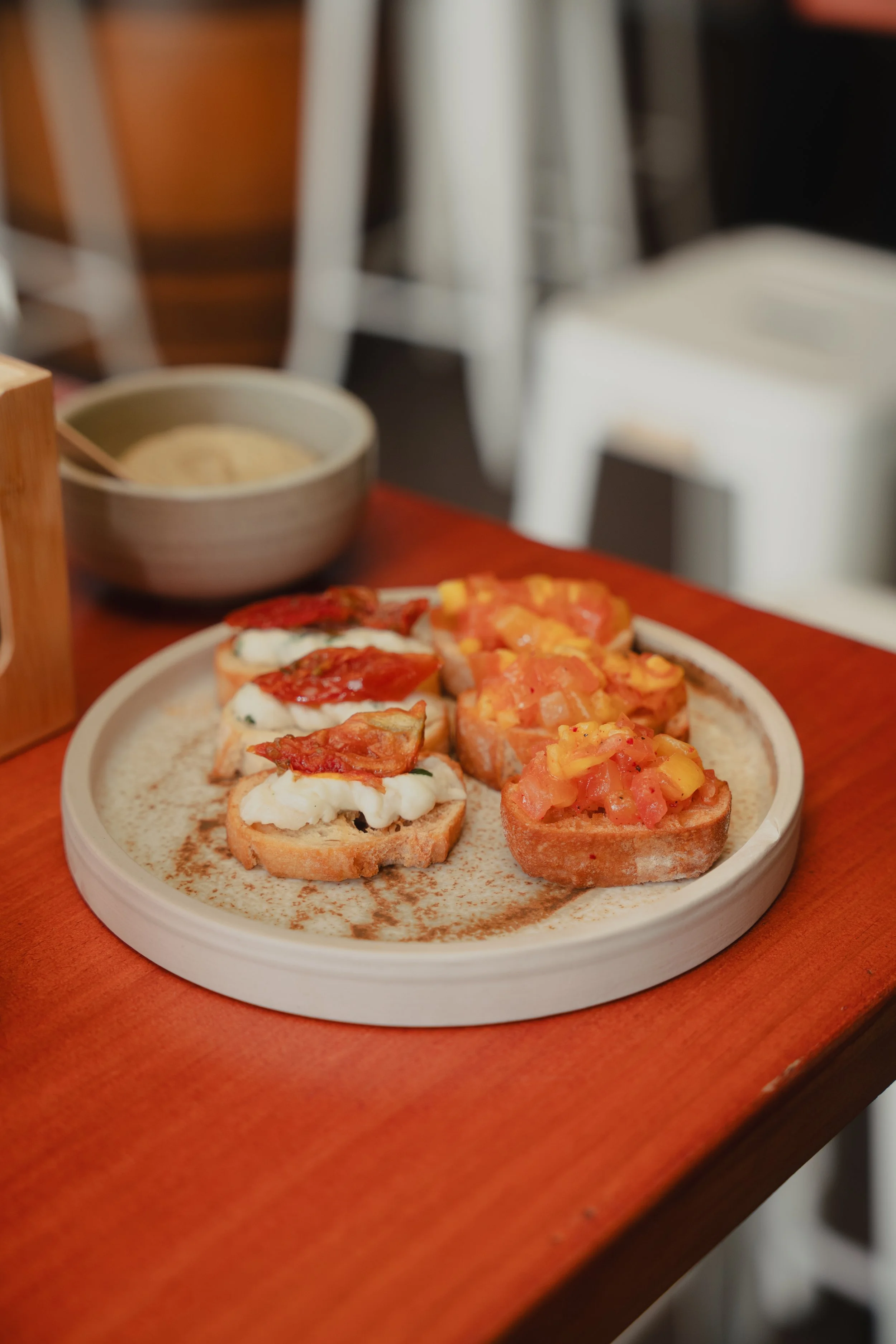 Close-up of a round plate with colorful bruschetta appetizers topped with chopped tomatoes, cheese, and herbs, placed on a wooden table. In the background, a bowl with dipping sauce and blurred chairs are visible.