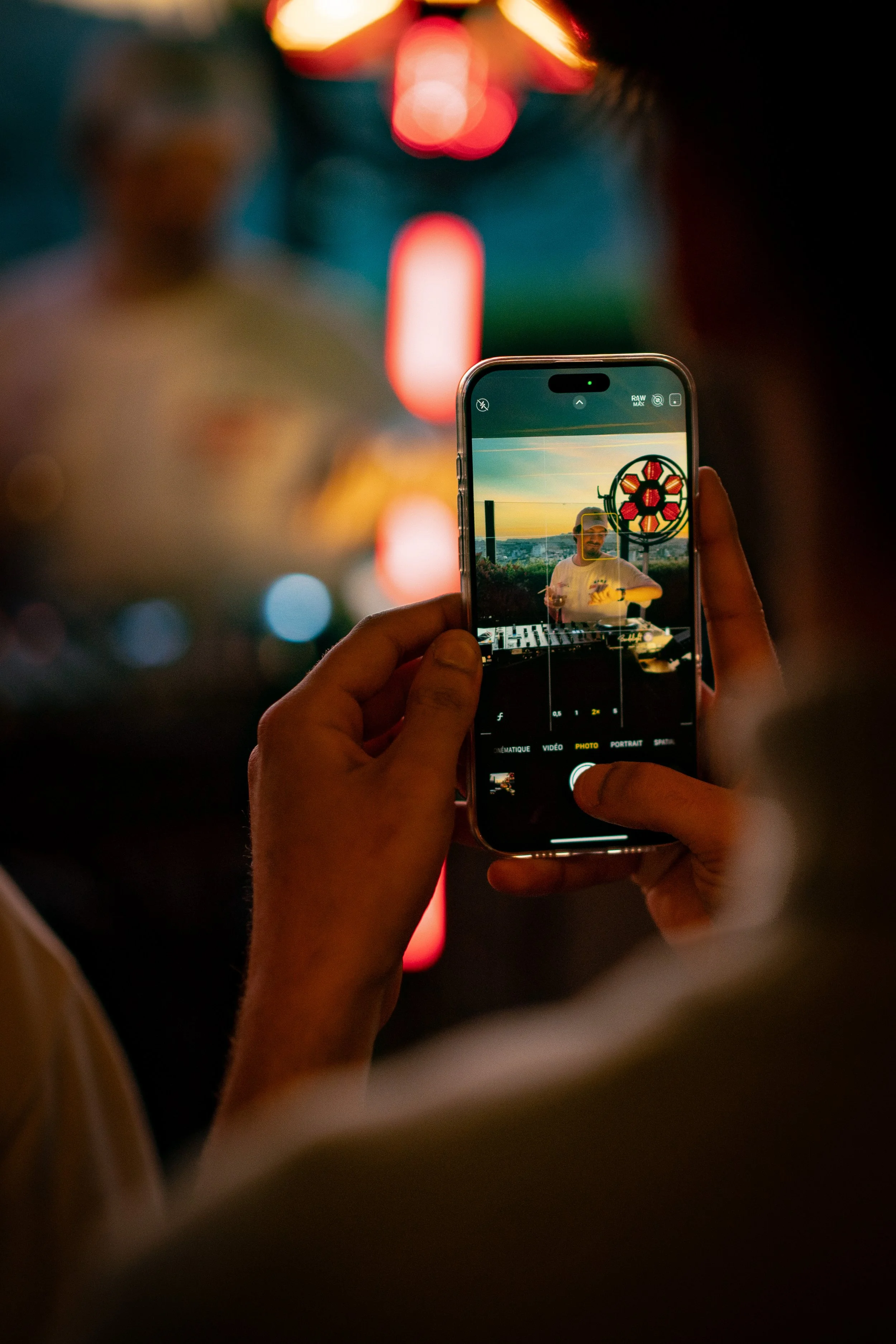 A person is using a smartphone to take a photo of a DJ at sunset, with the sky in the background.