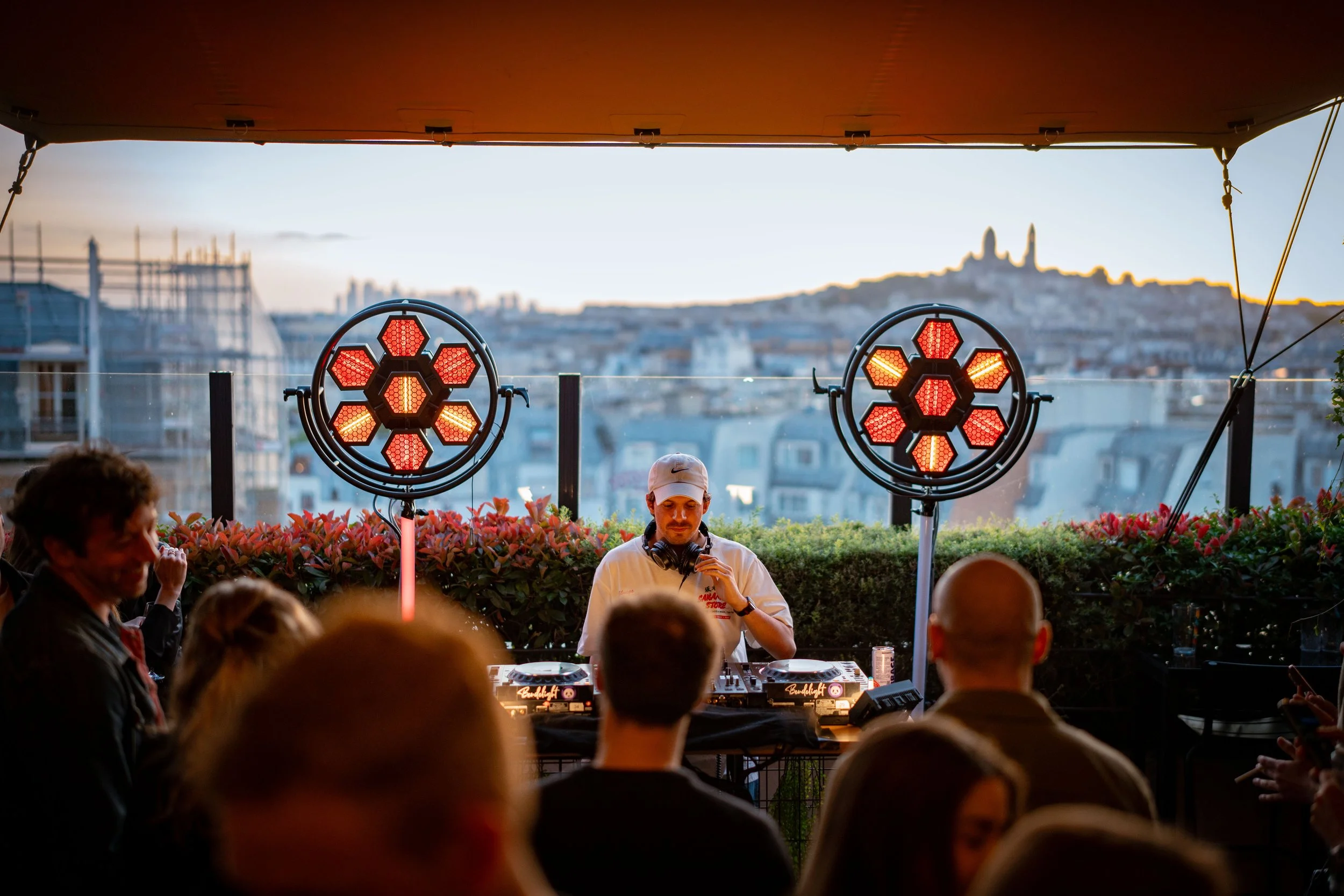A DJ wearing a white hoodie and headphones standing at a DJ booth during an outdoor event at sunset, illuminated by red hexagonal stage lights, with a crowd of people in the foreground and a cityscape in the background.