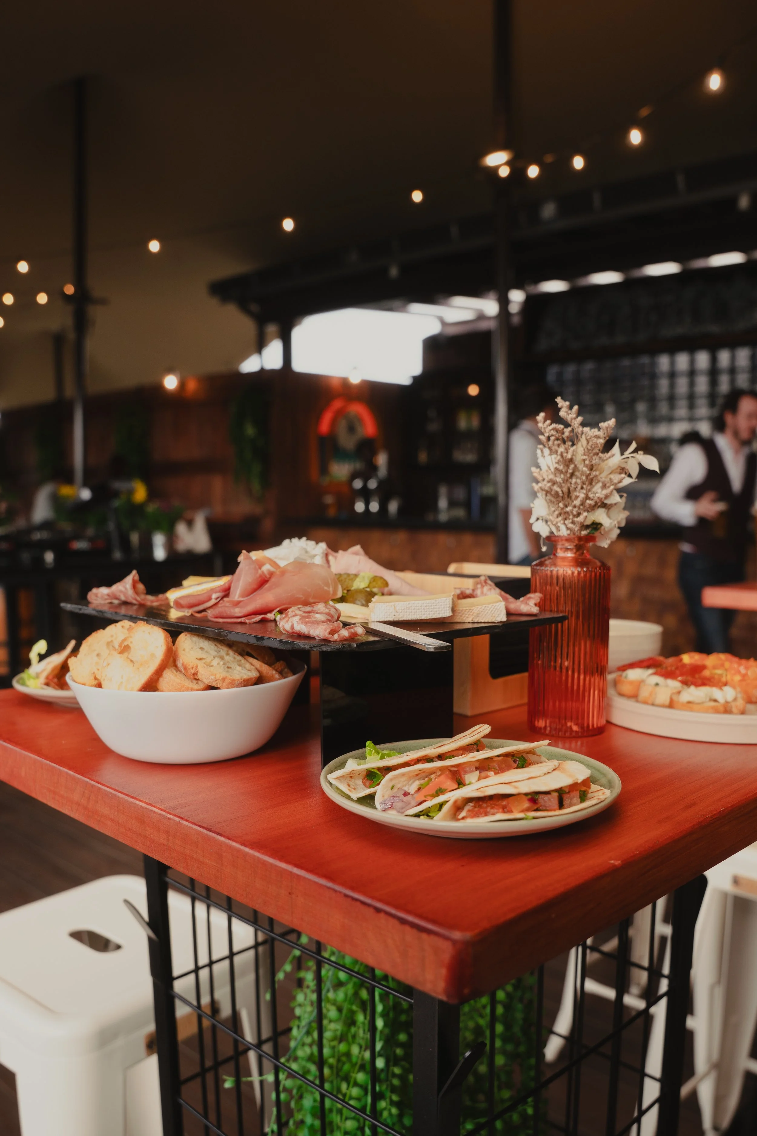 A close-up of a charcuterie and cheese platter on a red table, with bread slices and mini tacos in a white bowl. In the background, a person is standing in a cozy restaurant with warm lighting and wooden decor, and a vase with dried flowers on the ta