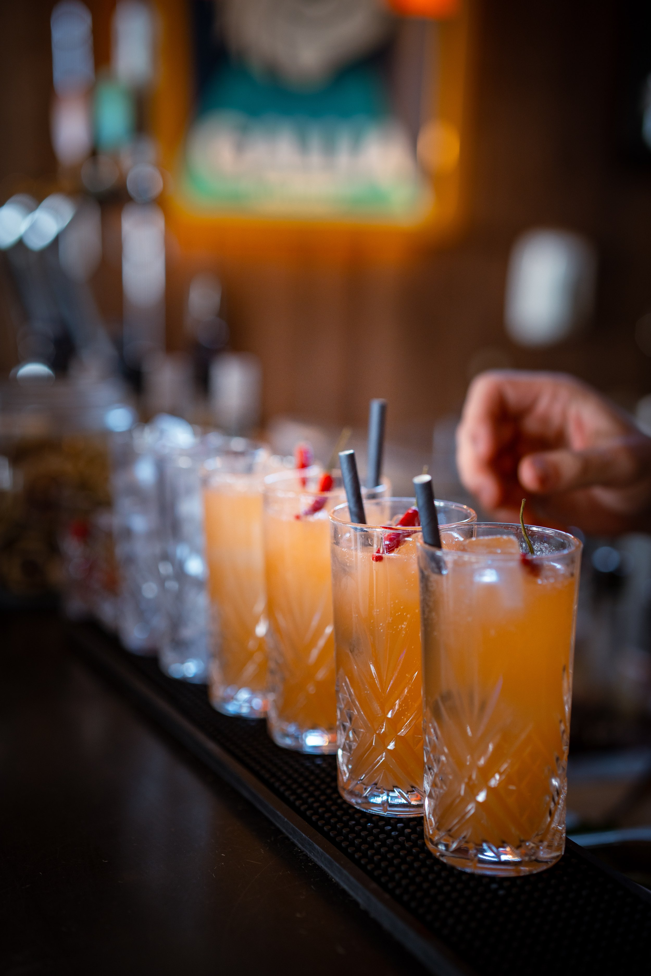 A row of peach-colored cocktails in ornate glassware with cherries and straws, sitting on a bar counter, with a blurred background of a poster and bar equipment.