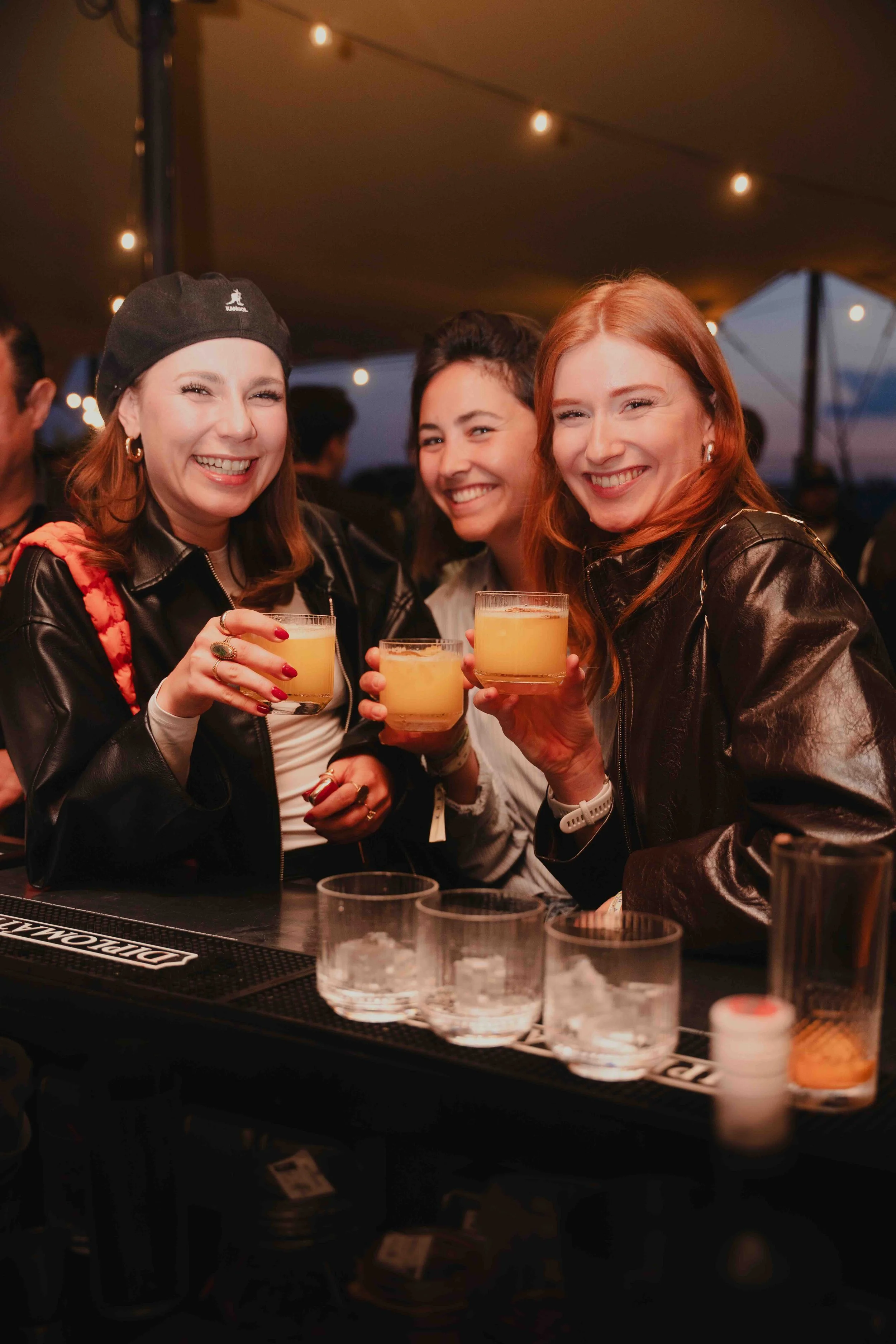 Three women smiling, holding drinks, standing behind a bar with shot glasses and bottles, under string lights at night.
