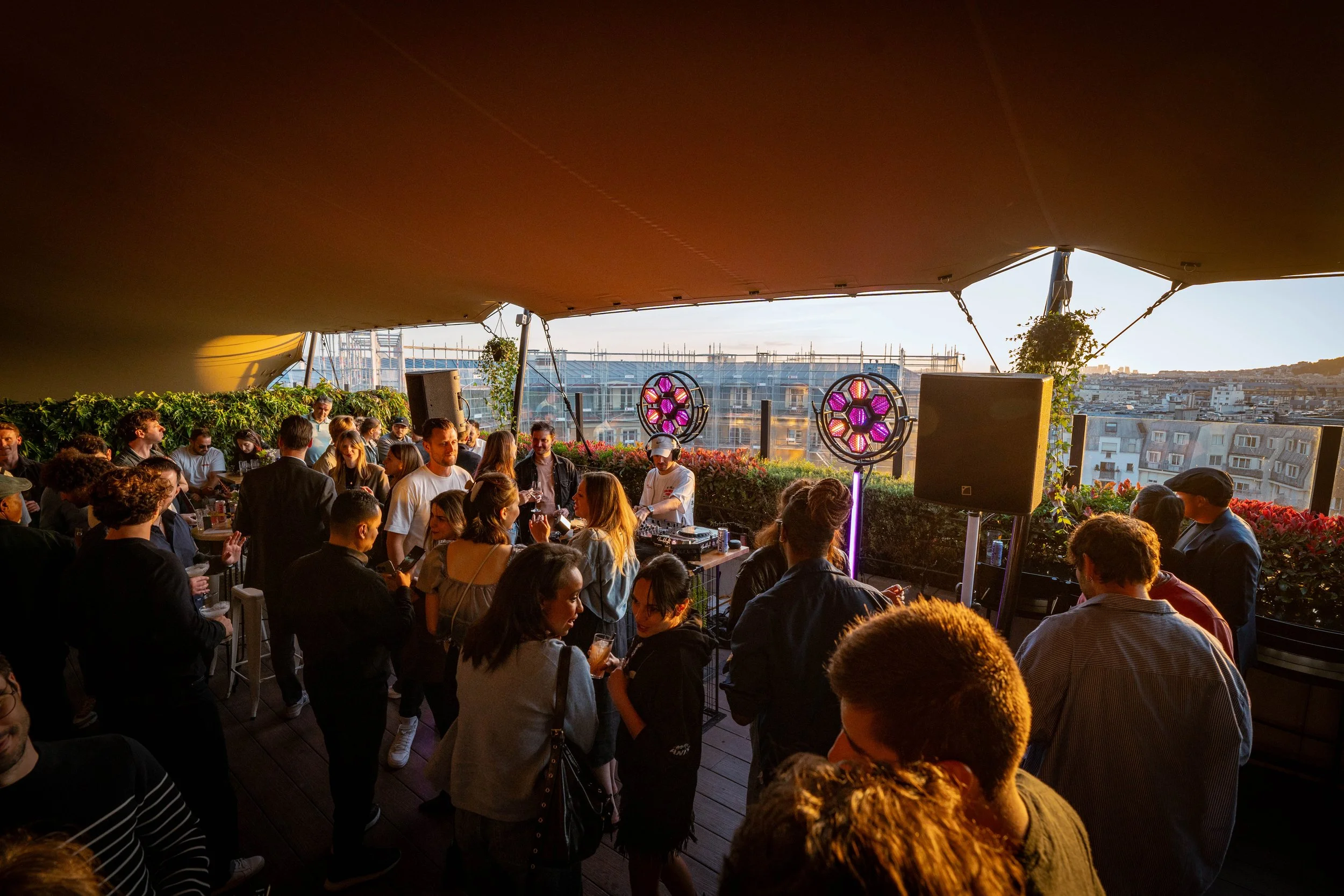 A rooftop gathering with people socializing and enjoying drinks at sunset, with cityscape in the background and colorful stage lighting.
