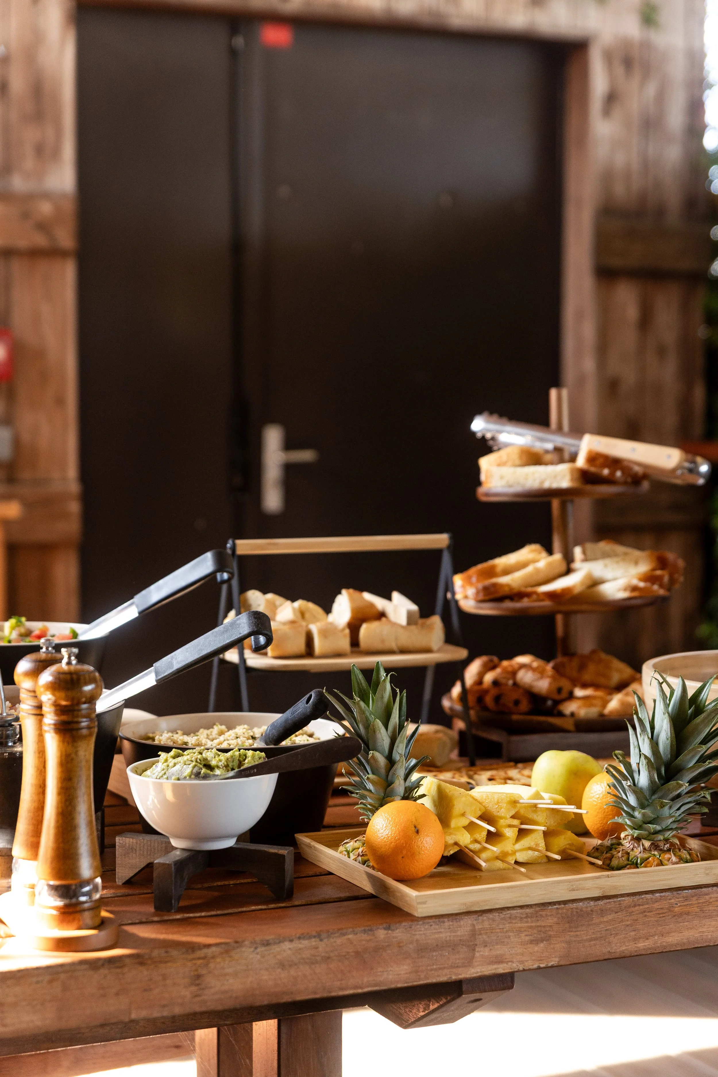 A wooden table set with a variety of foods including pineapples, oranges, skewers, and bowls of dips and guacamole. There is a multi-tiered tray with bread and baked goods, and a small bowl of chopped vegetables. The background features a dark door, part of a wooden wall, and some decorative items.