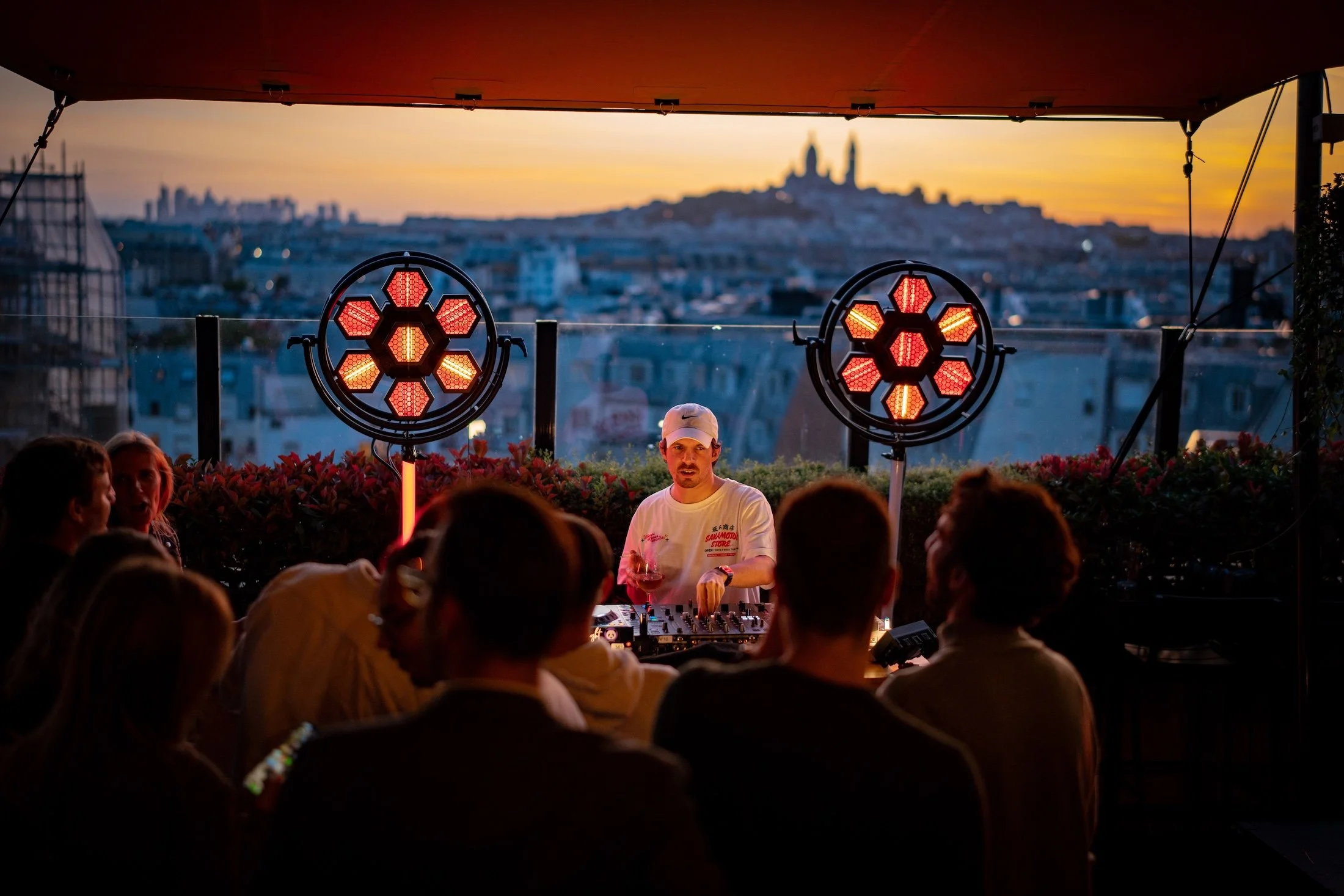 A DJ performing at an outdoor rooftop event during sunset with cityscape in the background, featuring two large red circular lights with honeycomb pattern.