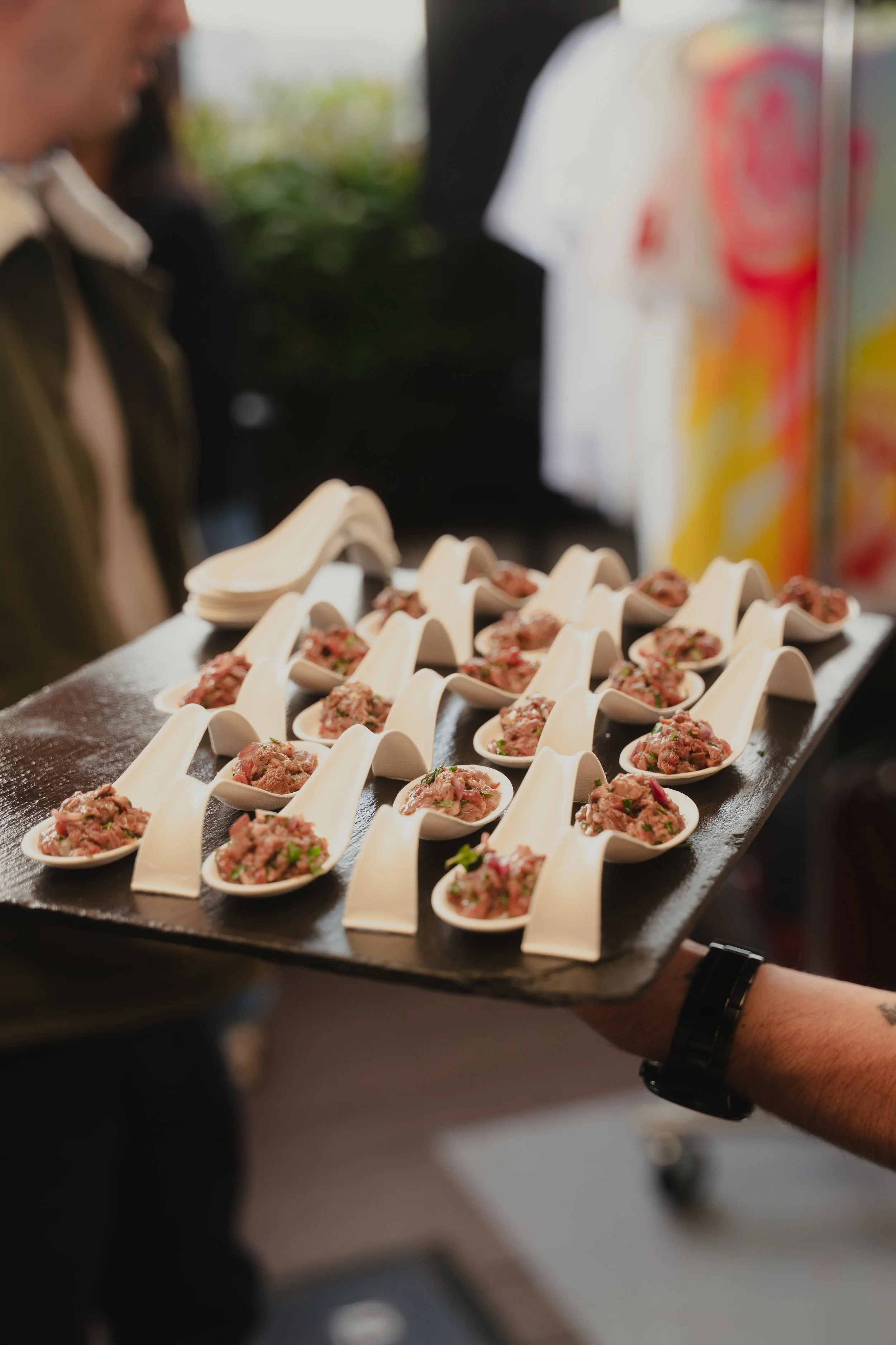 A person is holding a tray of small white spoons filled with raw minced meat garnished with herbs and spices, likely prepared for serving or cooking.