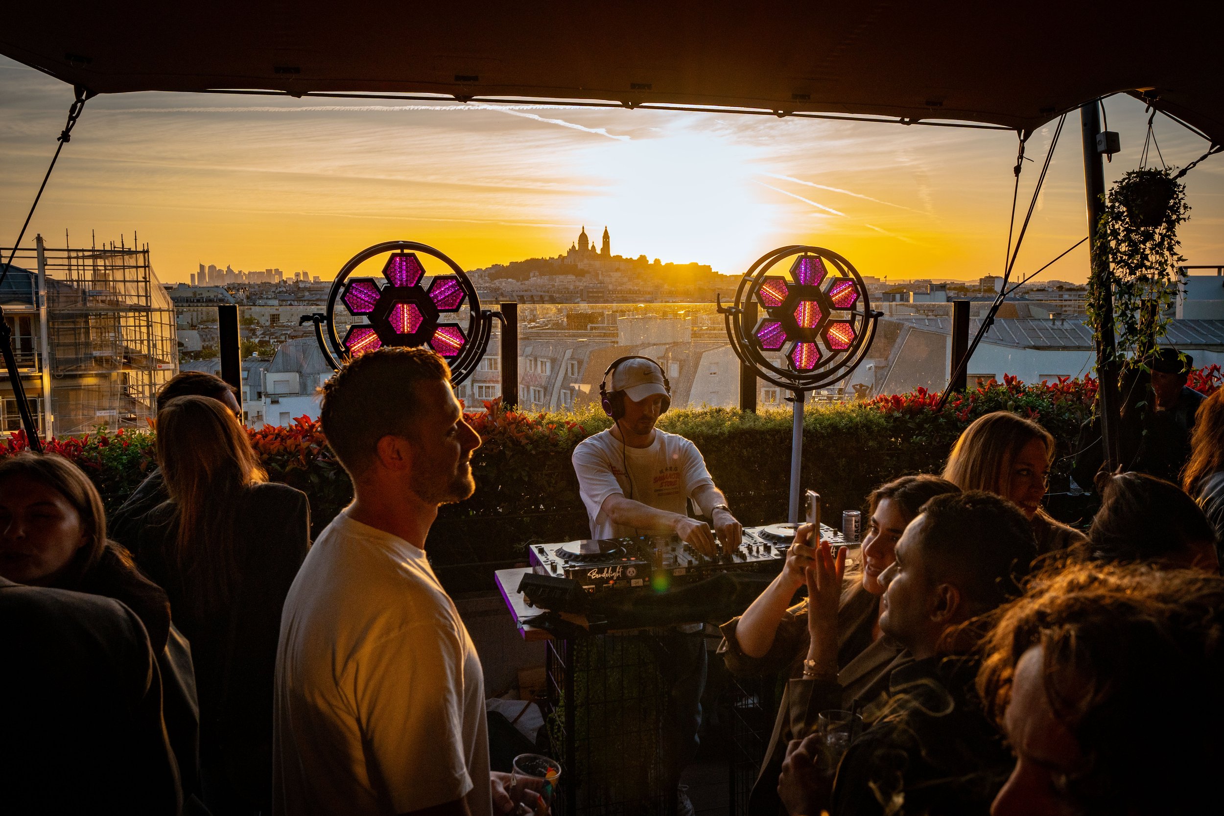 A rooftop party at sunset with a DJ playing music, surrounded by people socializing, with city buildings and a hilltop church in the background.