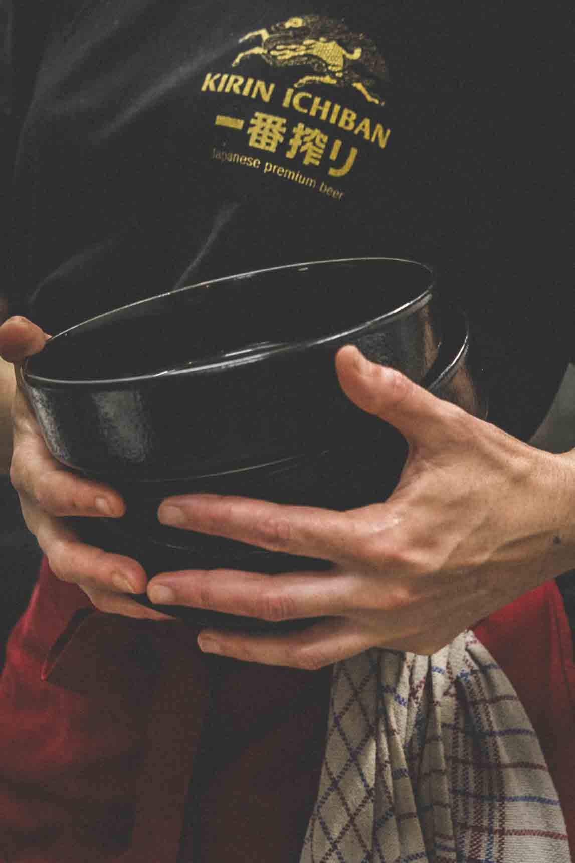 Personne tenant un bol noir, portant un t-shirt noir avec un logo et du texte en jaune