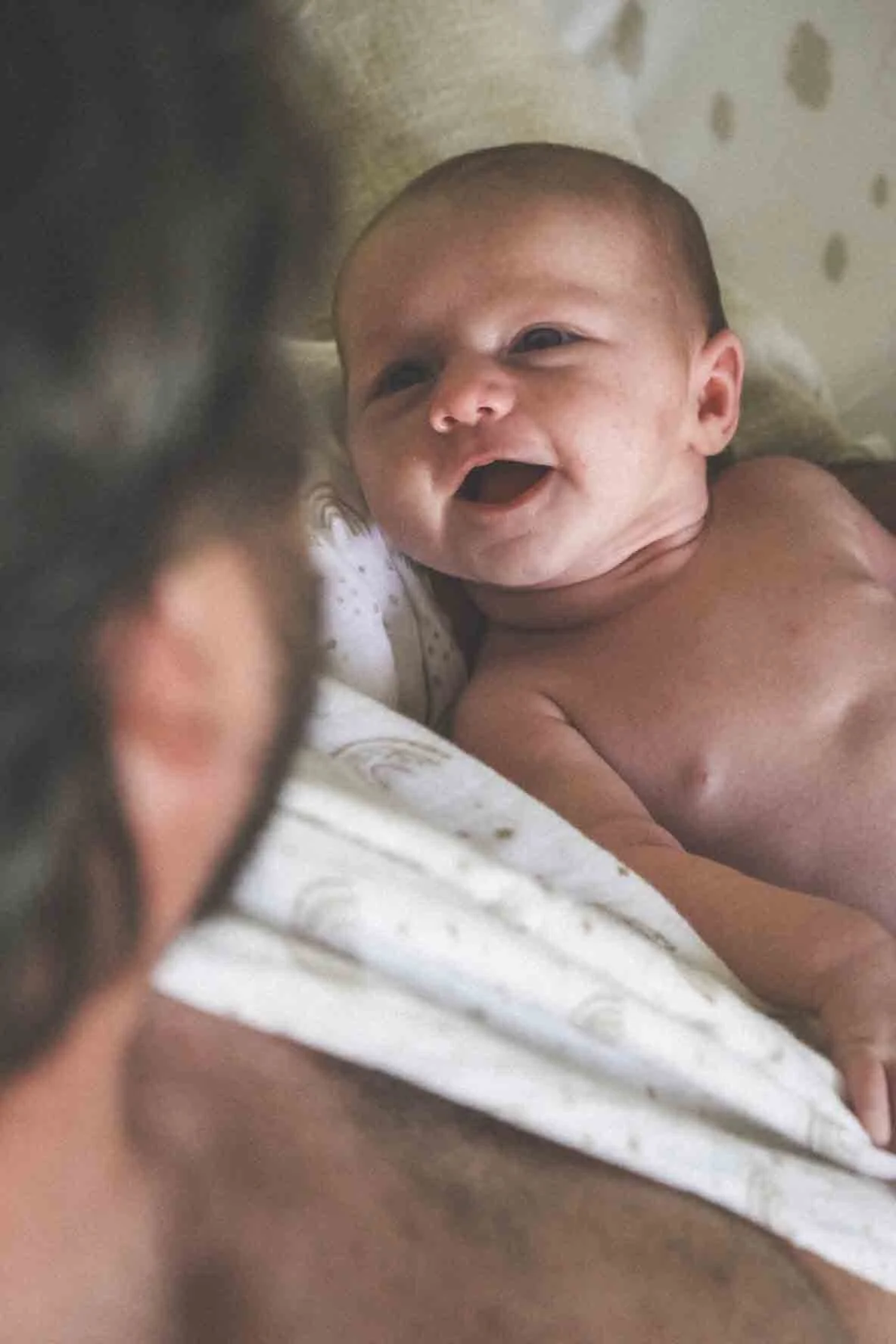 Un bébé souriant, allongé sur un lit, vu de profil avec un adulte dont seul une partie de la tête est visible en premier plan.