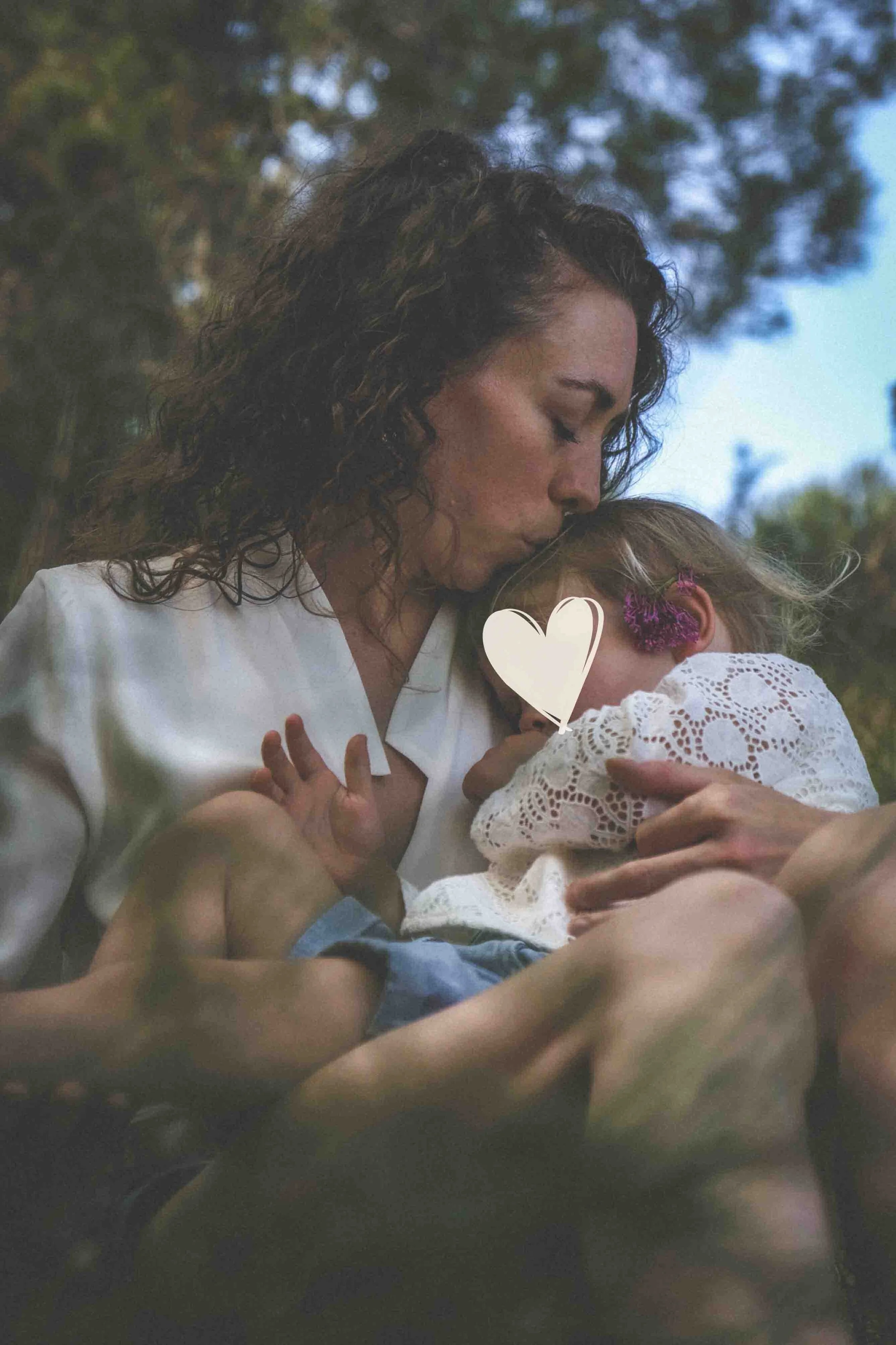 Une femme avec des cheveux bouclés embrasse un enfant dont elle tient la tête contre son épaule, la scène se déroule à l'extérieur avec des arbres en arrière-plan.