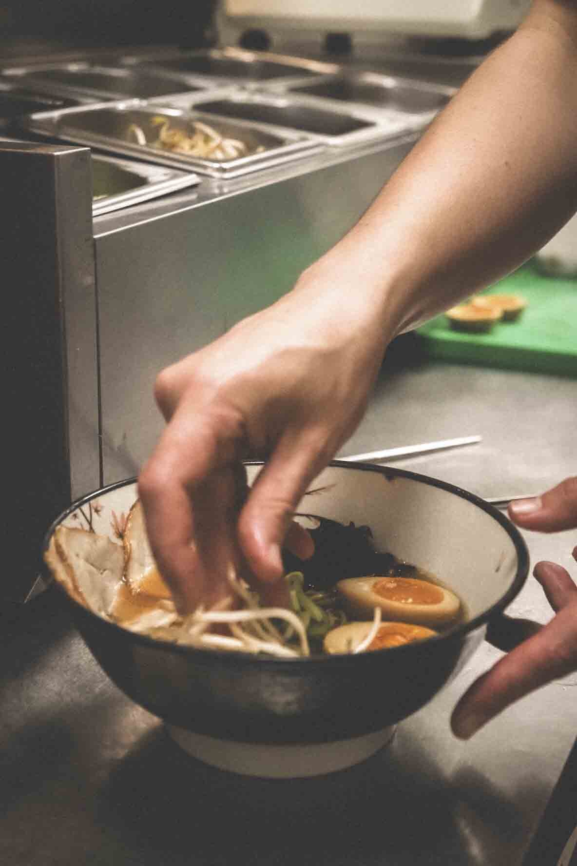 Main en train de préparer un bol de ramen avec œuf à moitié cuit, nouilles, légumes et viande, dans une cuisine.