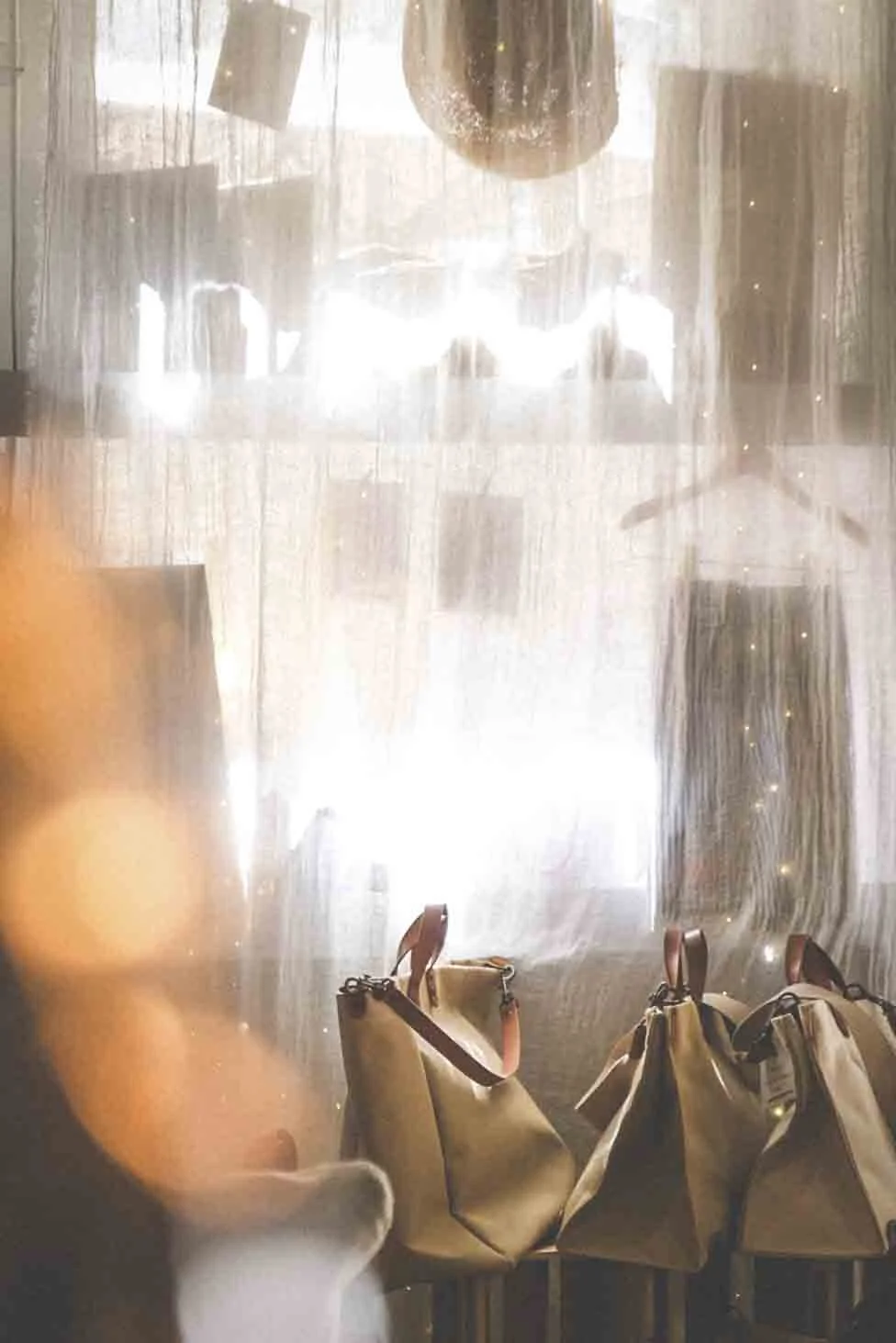 Vus à travers une fenêtre en verre dépolie ou une porte vitrée, plusieurs sacs en tissu ou en cuir posés sur des tabourets. La lumière provient d'une source extérieure ou intérieure, créant des reflets brillants sur la surface en verre.