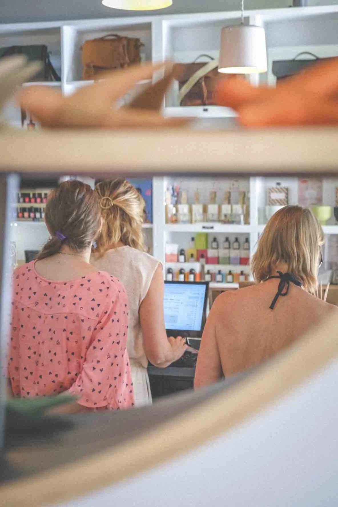 Trois femmes travaillent sur un ordinateur dans une boutique ou un salon, vue partielle à travers une étagère avec des sacs en cuir et des lampes en arrière-plan.