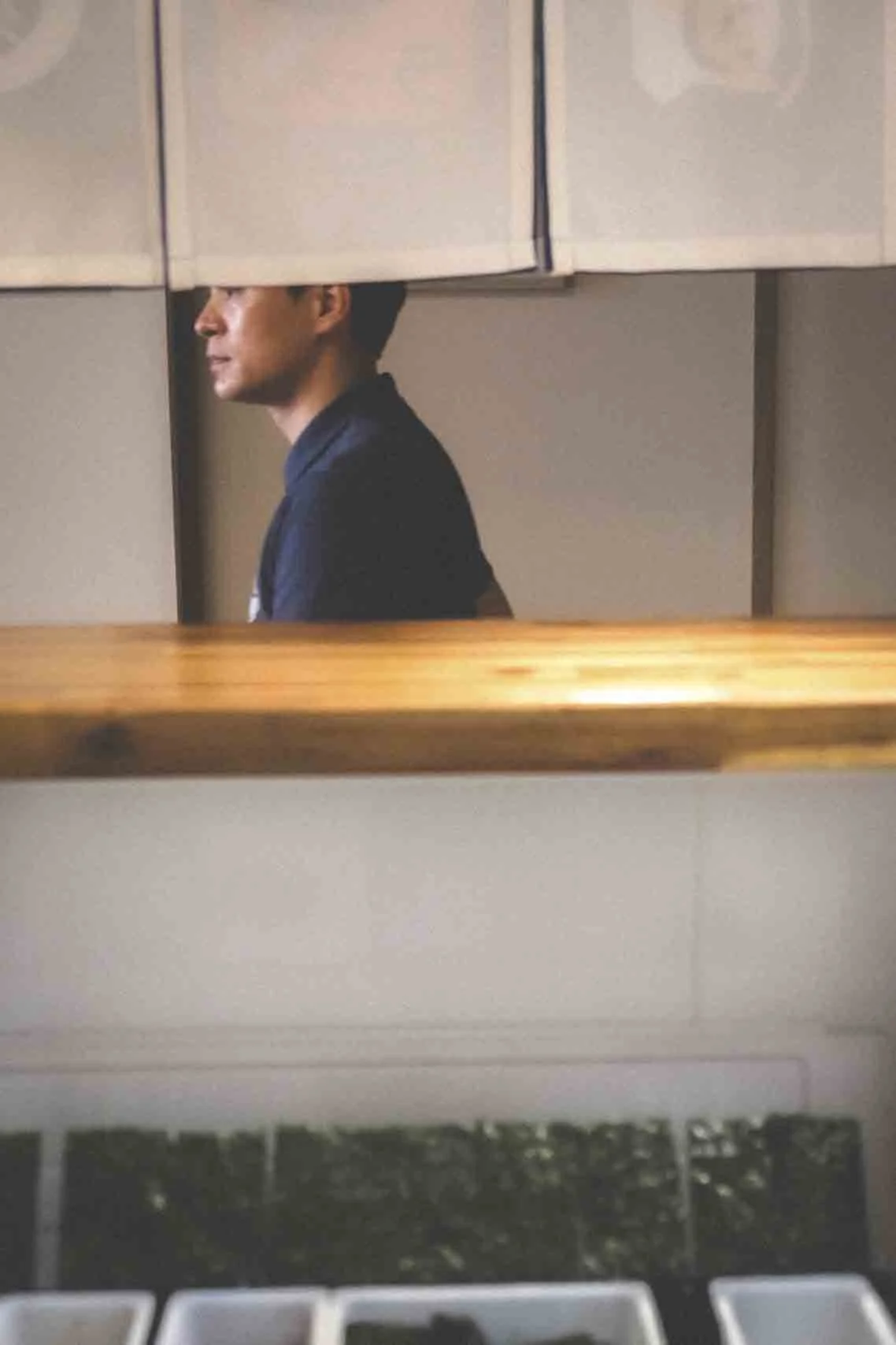 Un homme vêtu d'une chemise sombre regarde vers la gauche, vu à travers un miroir dans une salle propre et moderne, avec un comptoir en bois au premier plan. Le reflet montre un intérieur bien éclairé.