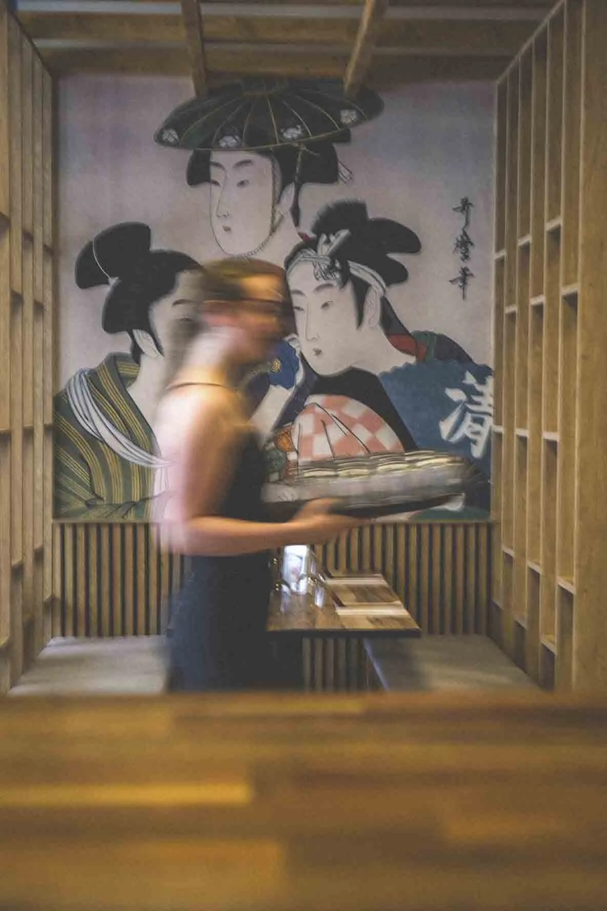 Une femme en noir tenant un plateau dans un restaurant avec un mur décoratif japonais en arrière-plan, représentant des geishas traditionnelles.