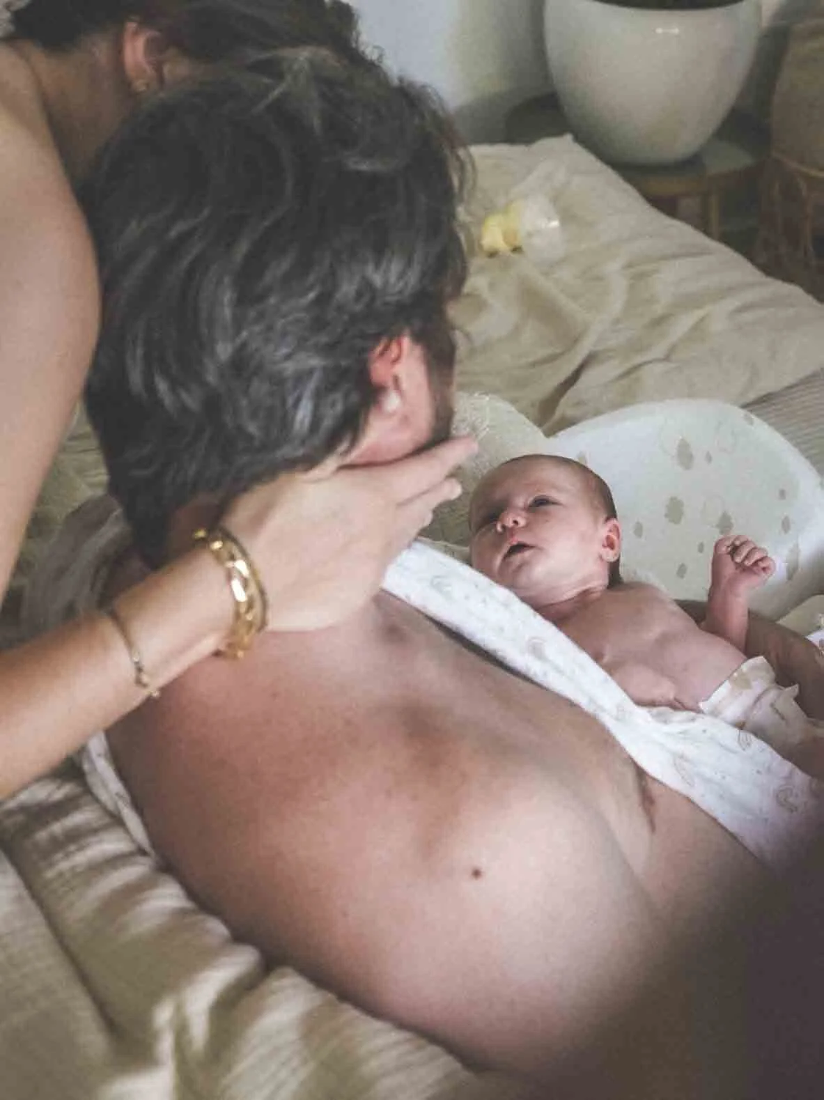 Une mère et un père regardent un nouveau-né dans une pièce. La mère porte des bijoux en or et regarde tendrement le bébé, qui est allongé dans un berceau. La scène semble se passer dans une maison, avec un lit et un décor simple en arrière-plan.