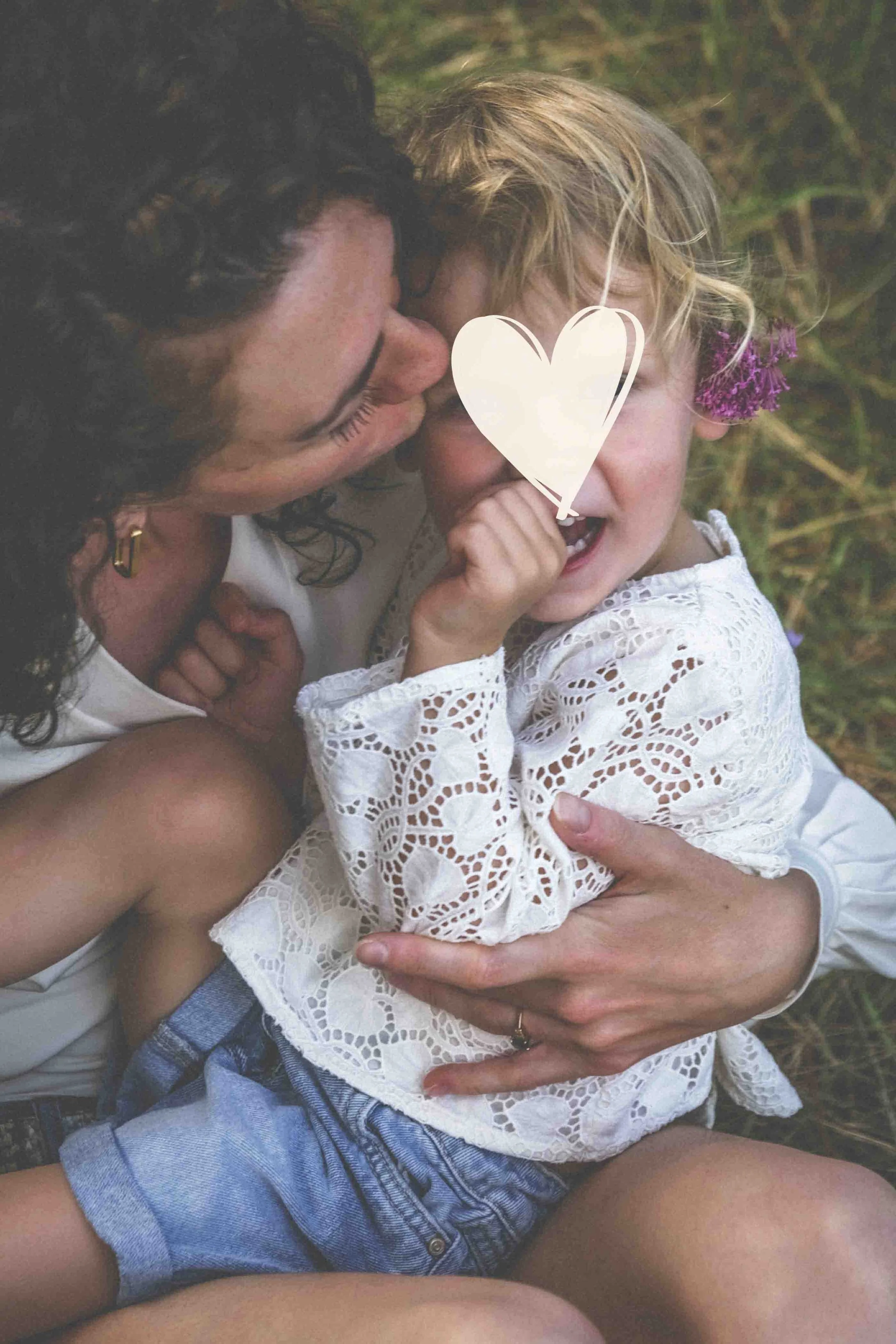 Une femme et une petite fille, probablement sa fille, sont allongées dans l'herbe. La femme embrasse tendrement la petite fille, qui porte une robe en dentelle blanche et a une fleur violette dans les cheveux, dans un contexte de plein air.